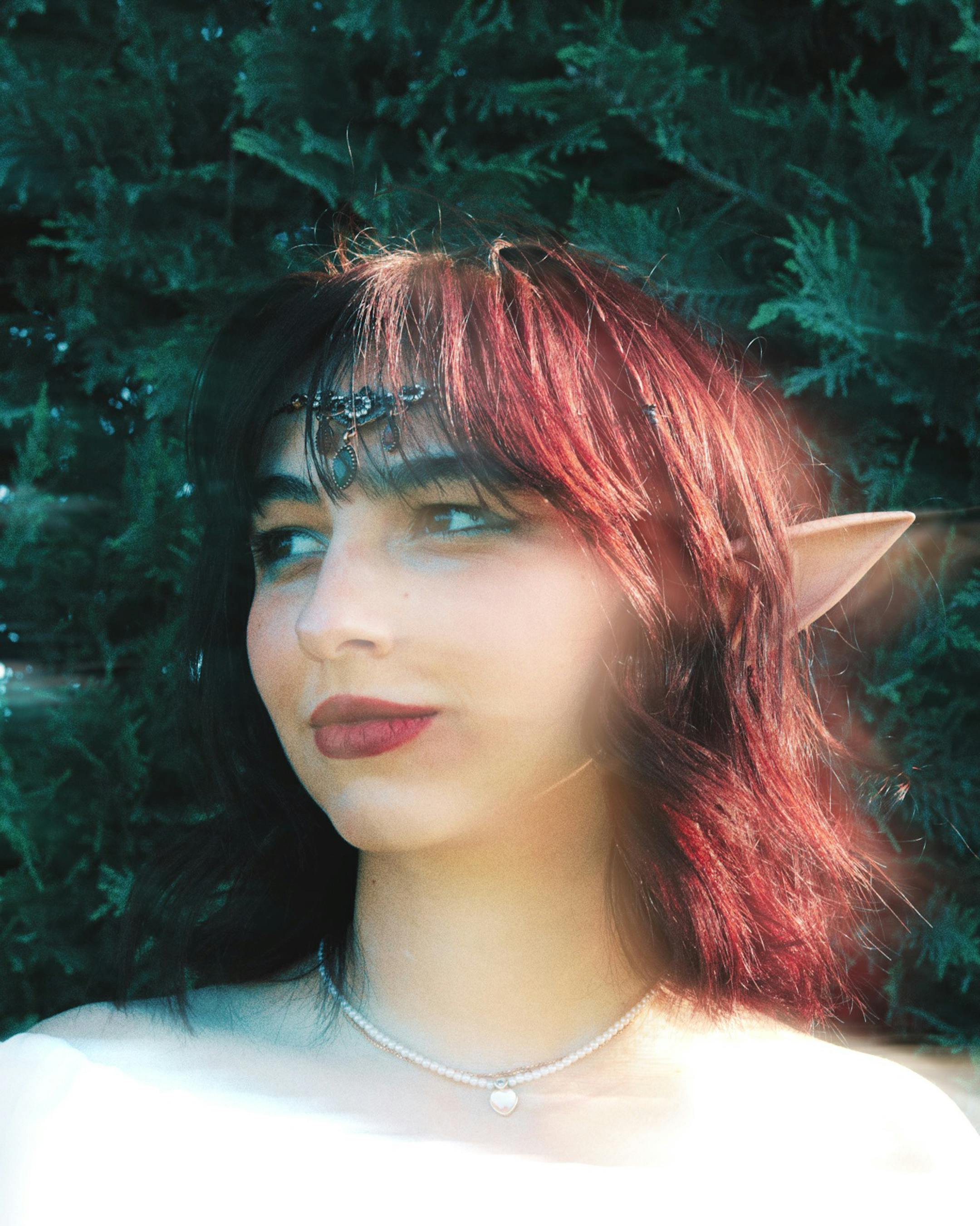 A fantasy-themed portrait of a woman with elf ears and dyed hair in a natural outdoor setting.