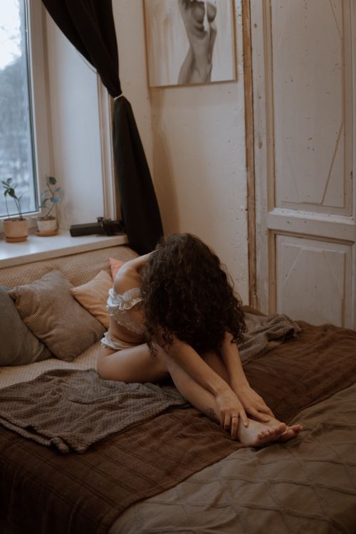 Free A woman sits on a bed with brown sheets in a cozy bedroom, creating a warm, contemplative atmosphere. Stock Photo