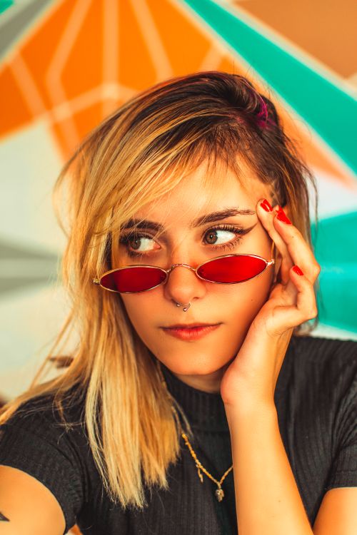 Free Trendy young woman posing with red sunglasses against a vibrant geometric background, showcasing contemporary style. Stock Photo
