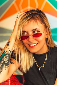 一位戴着红色太阳镜、留着金色头发的纹身女子，在五彩斑斓的背景下灿烂地微笑着