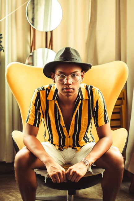 Fashionable man wearing yellow striped shirt and hat seated in a stylish chair indoors.