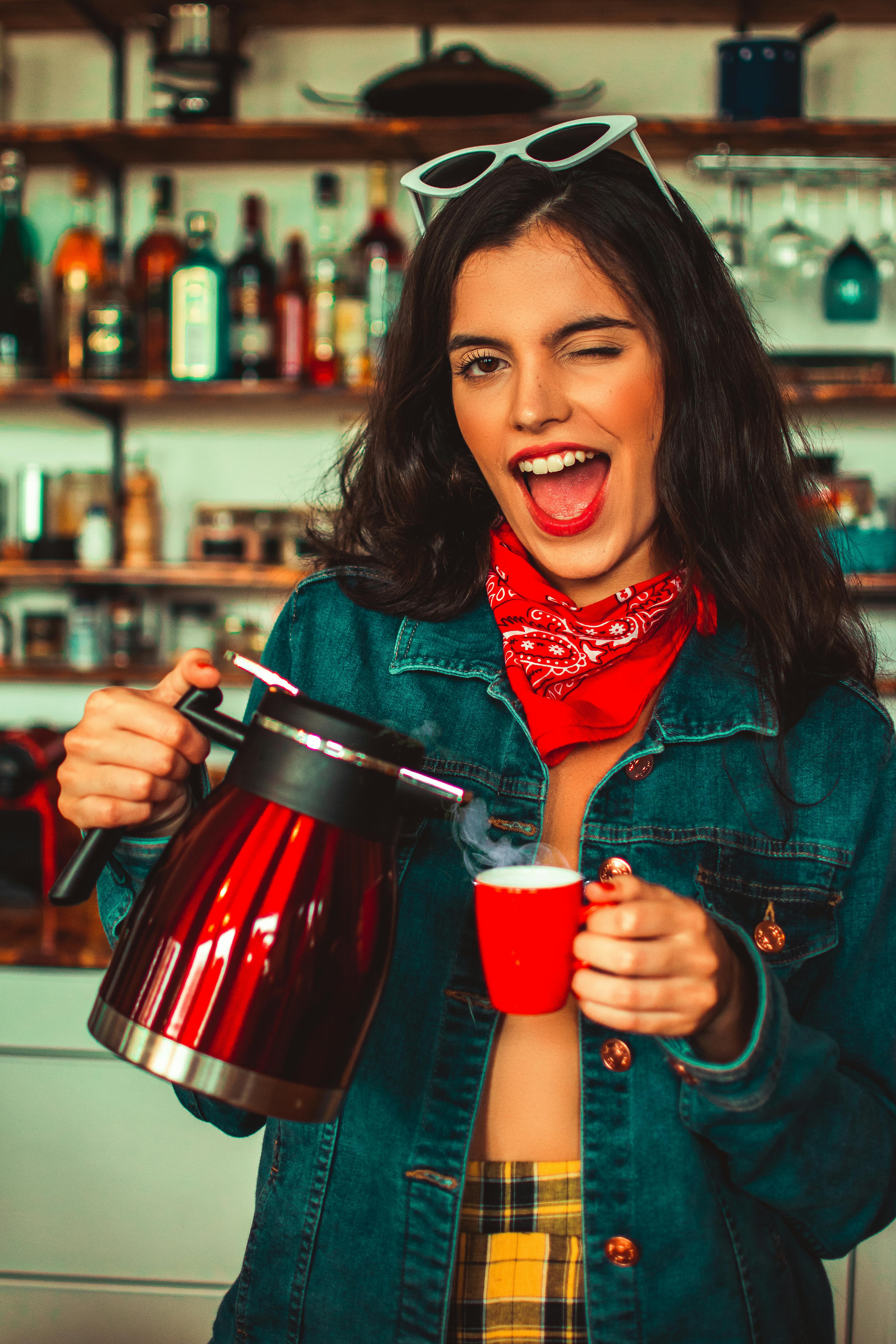 photo of winking woman in blue denim jacket holding red electric kettle and cup