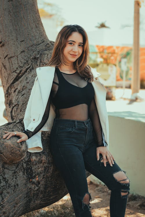 A woman in black top and ripped jeans leaning against a tree