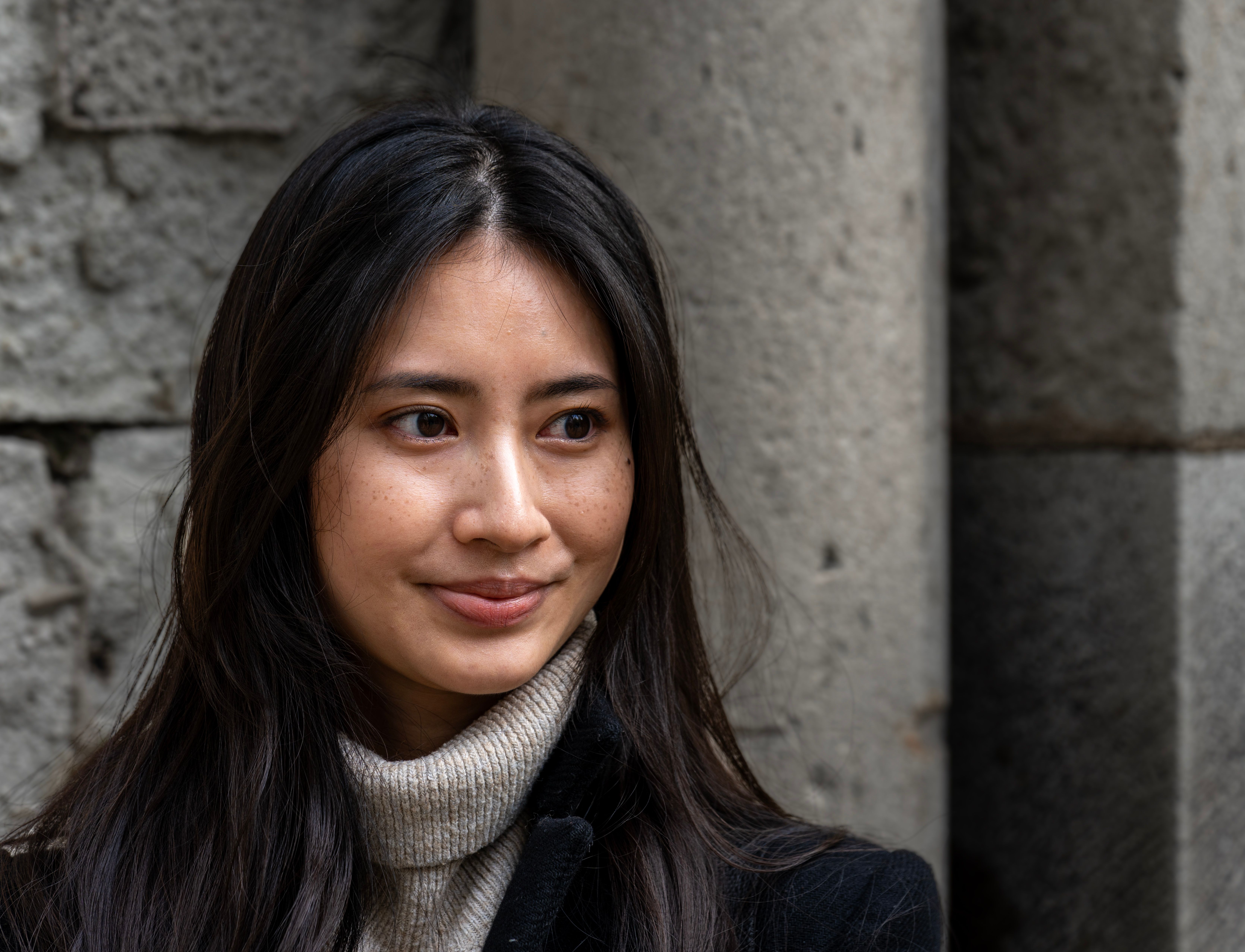 A woman with long black hair smiling softly in an urban setting with stone walls.