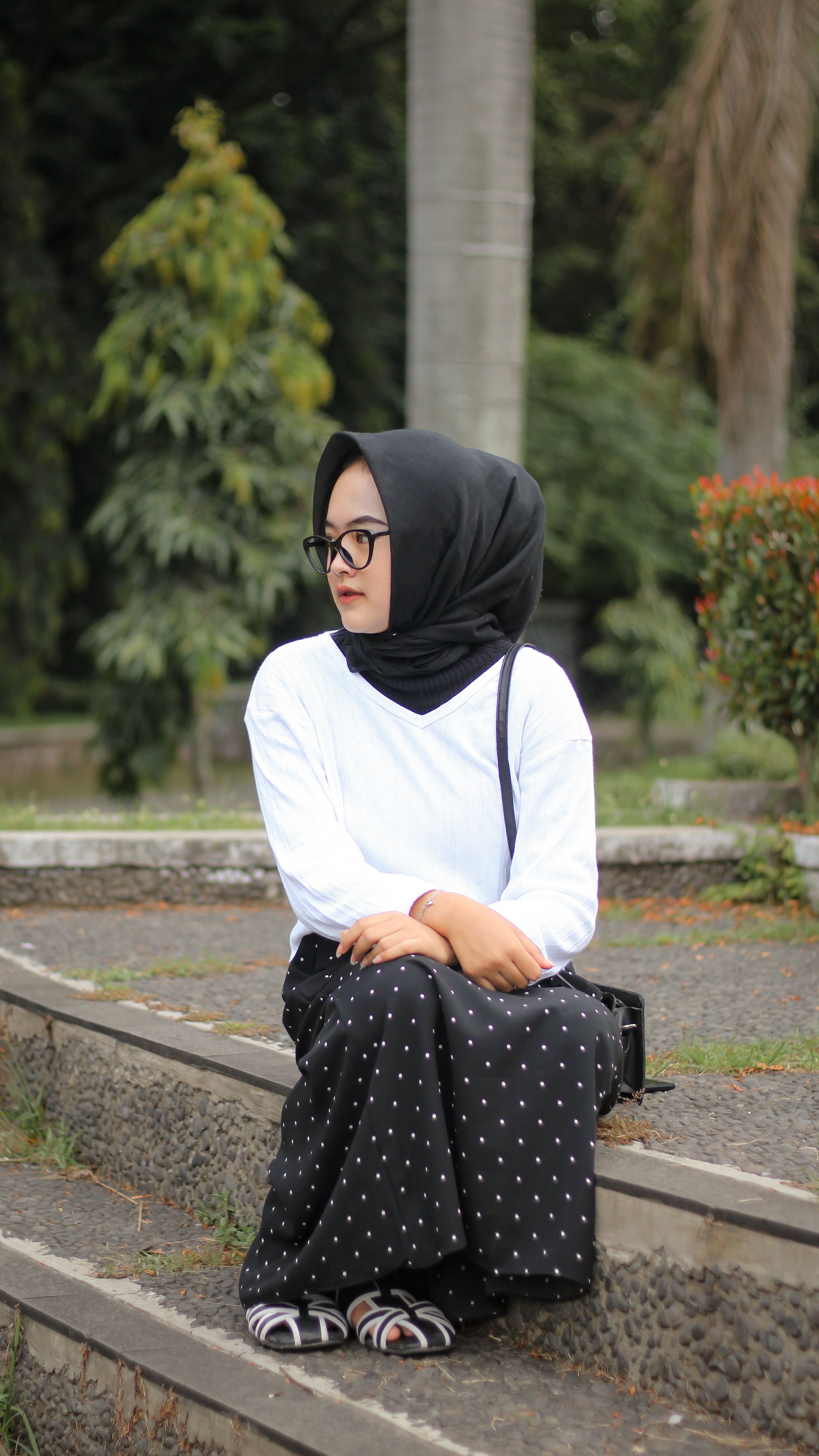 A woman wearing a black headscarf sits on stairs in a green park in Bandung, Indonesia.