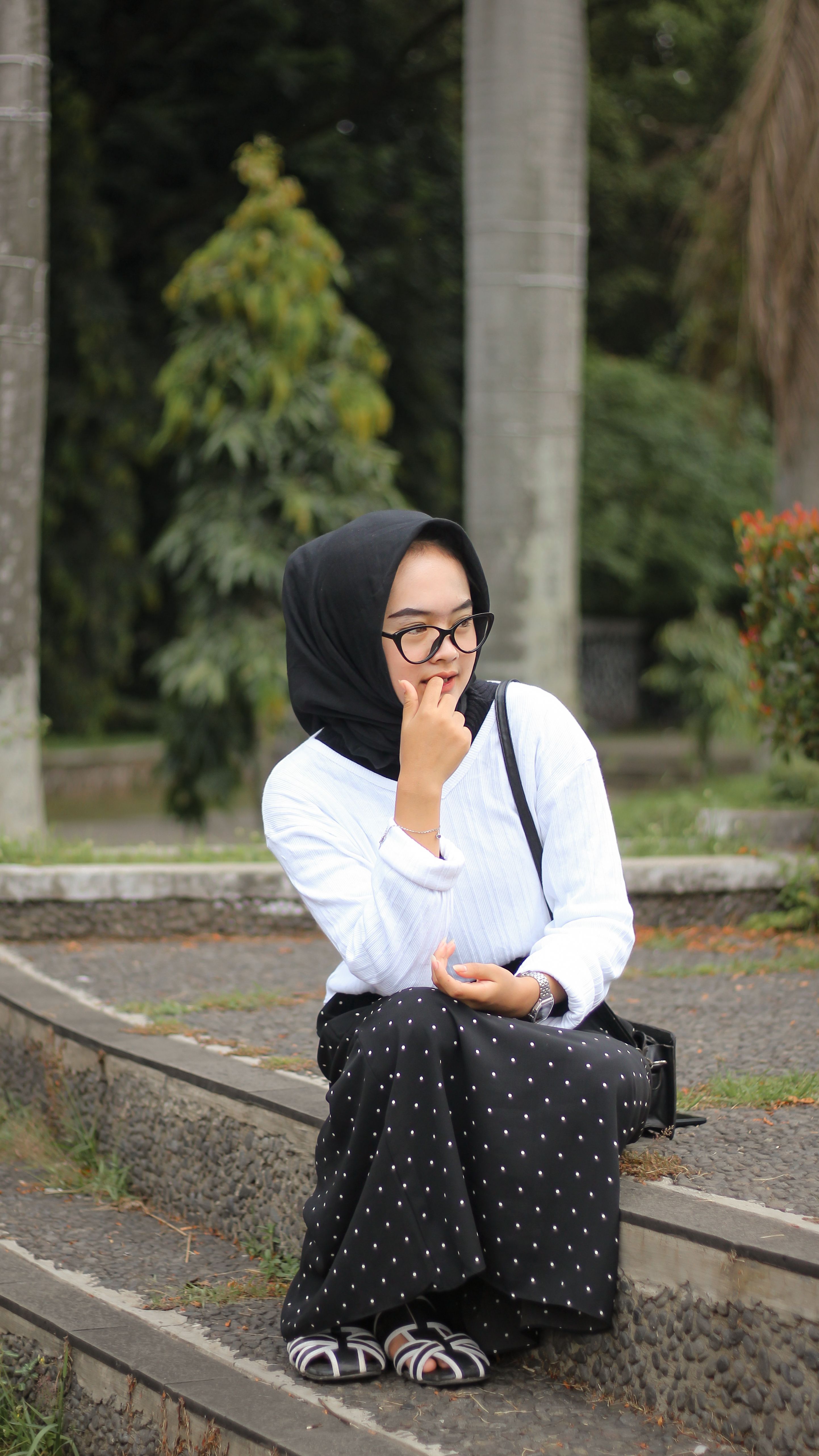 A young woman in a headscarf sits pensively on park stairs, surrounded by greenery.