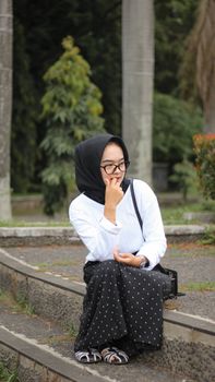 A young woman in a headscarf sits pensively on park stairs, surrounded by greenery. - Fotografer Sidoarjo