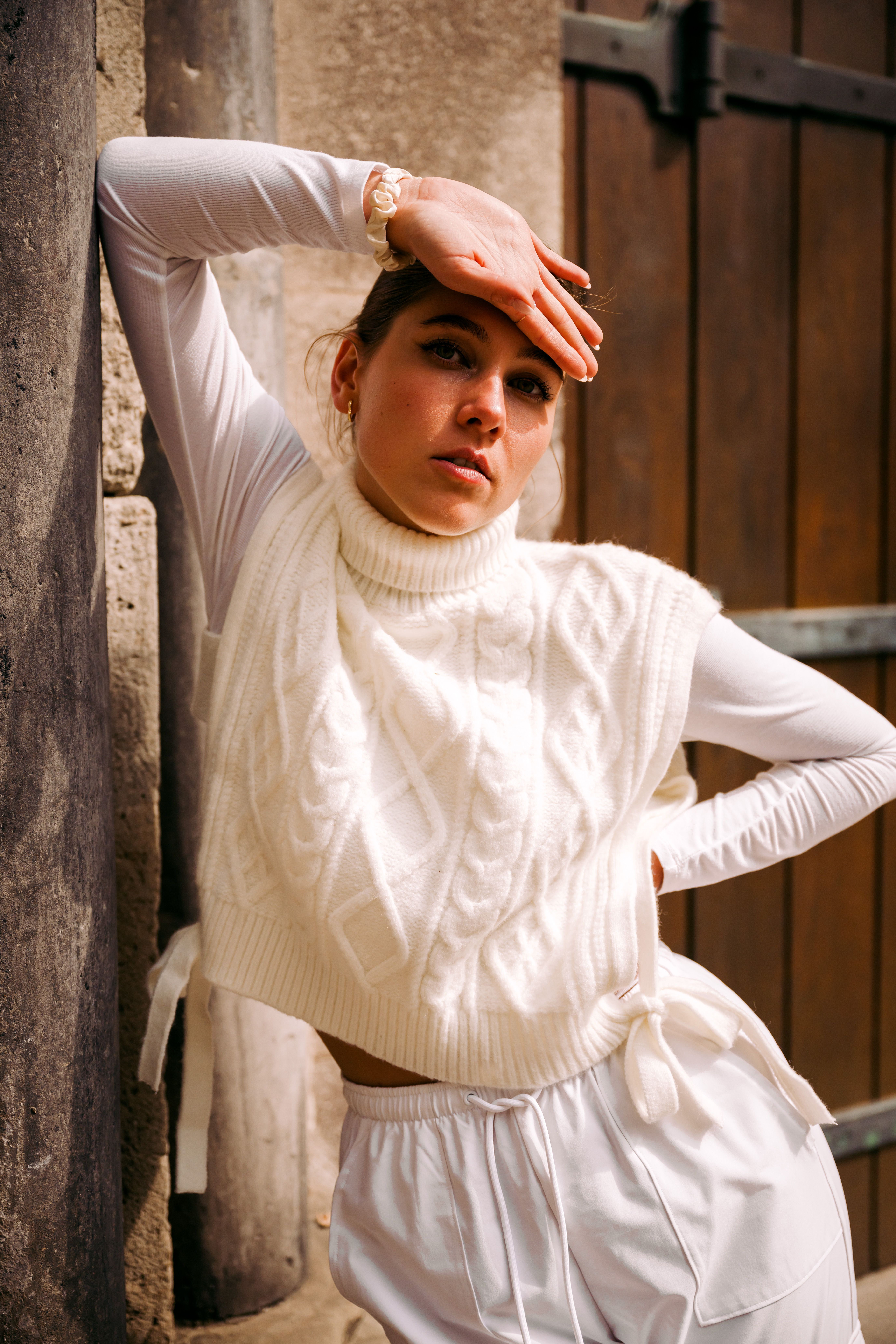 Stylish young woman in a cozy knitted turtleneck vest poses with elegance by a stone wall, outdoors.