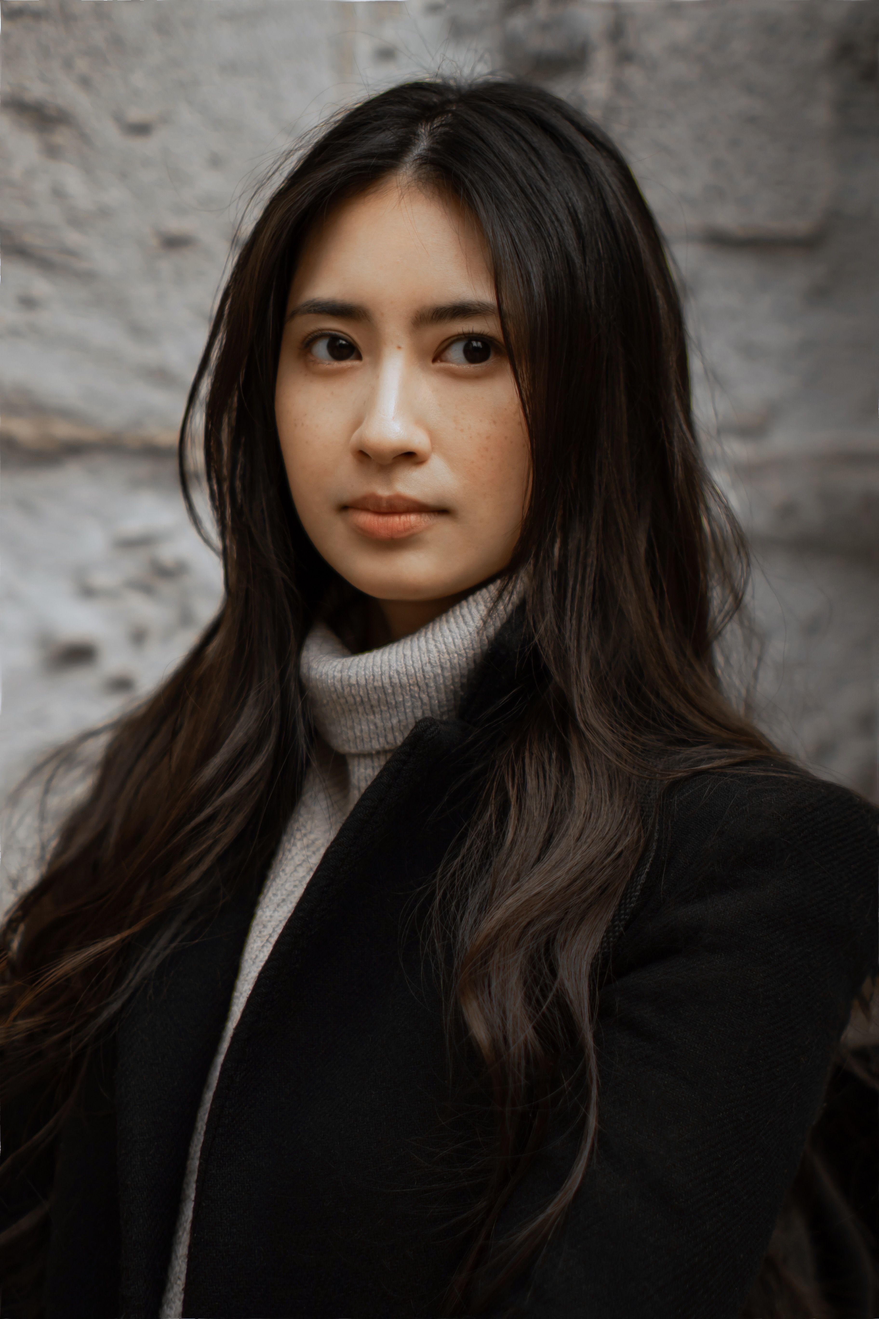 Close-up portrait of a young woman in a black jacket against a stone wall.