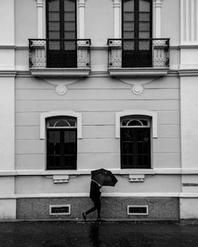 Black and white image of elegant building facade with a lone walker holding an umbrella.