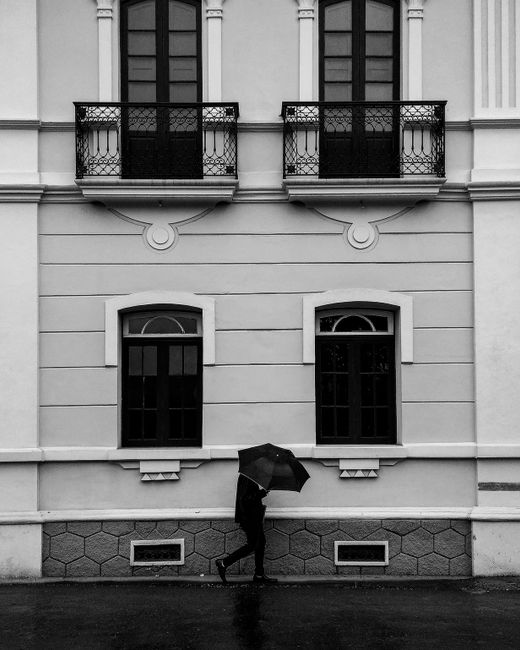 Black and white image of elegant building facade with a lone walker holding an umbrella.