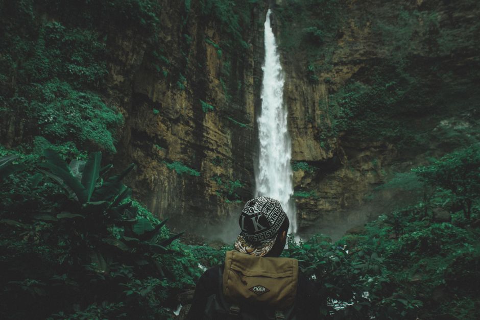 Person admiring a tall waterfall in a lush, green forest, conveying adventure and exploration.