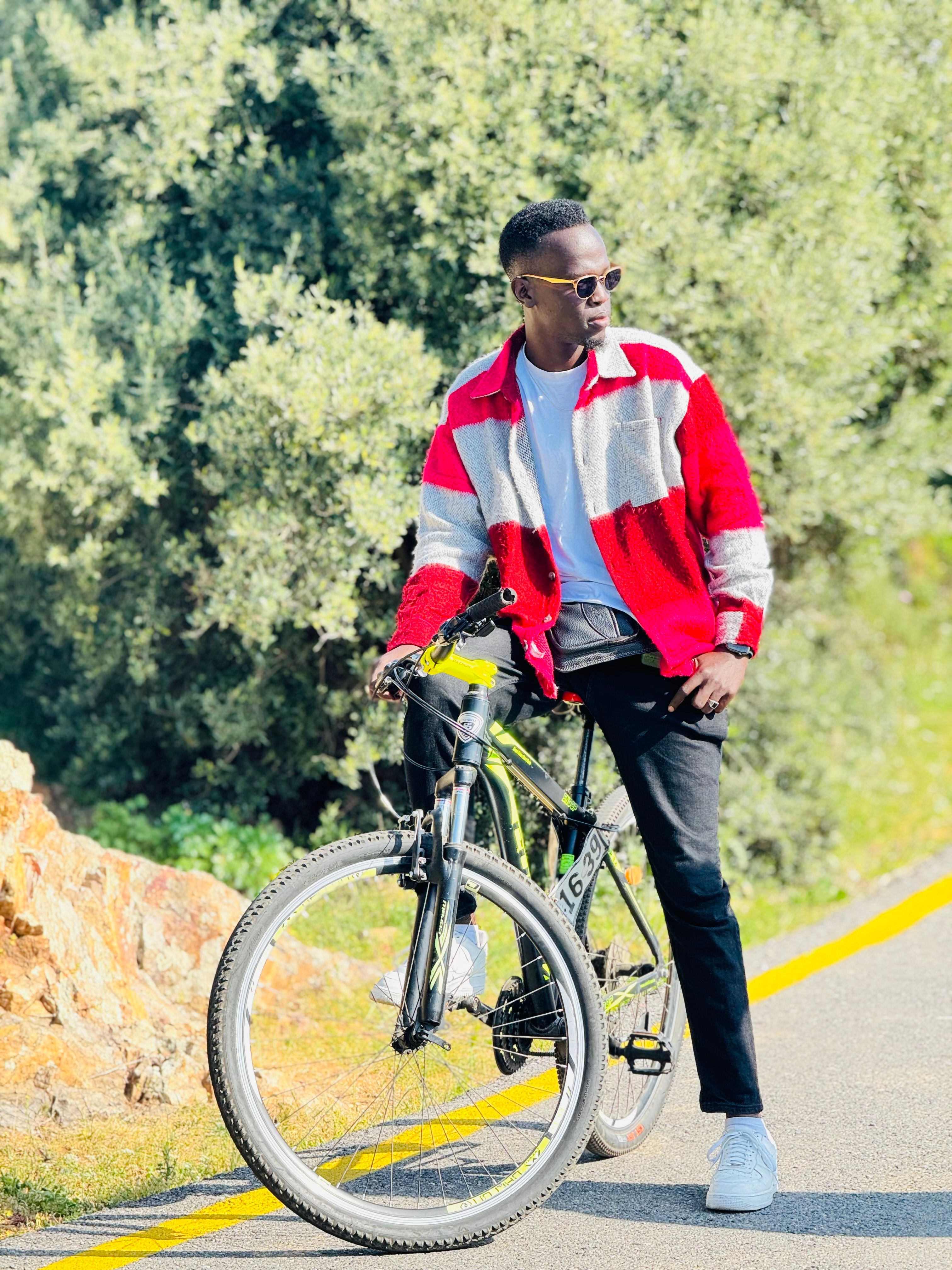 Model wearing red jacket, seated on a bicycle amidst greenery, exuding fashion and casual style.