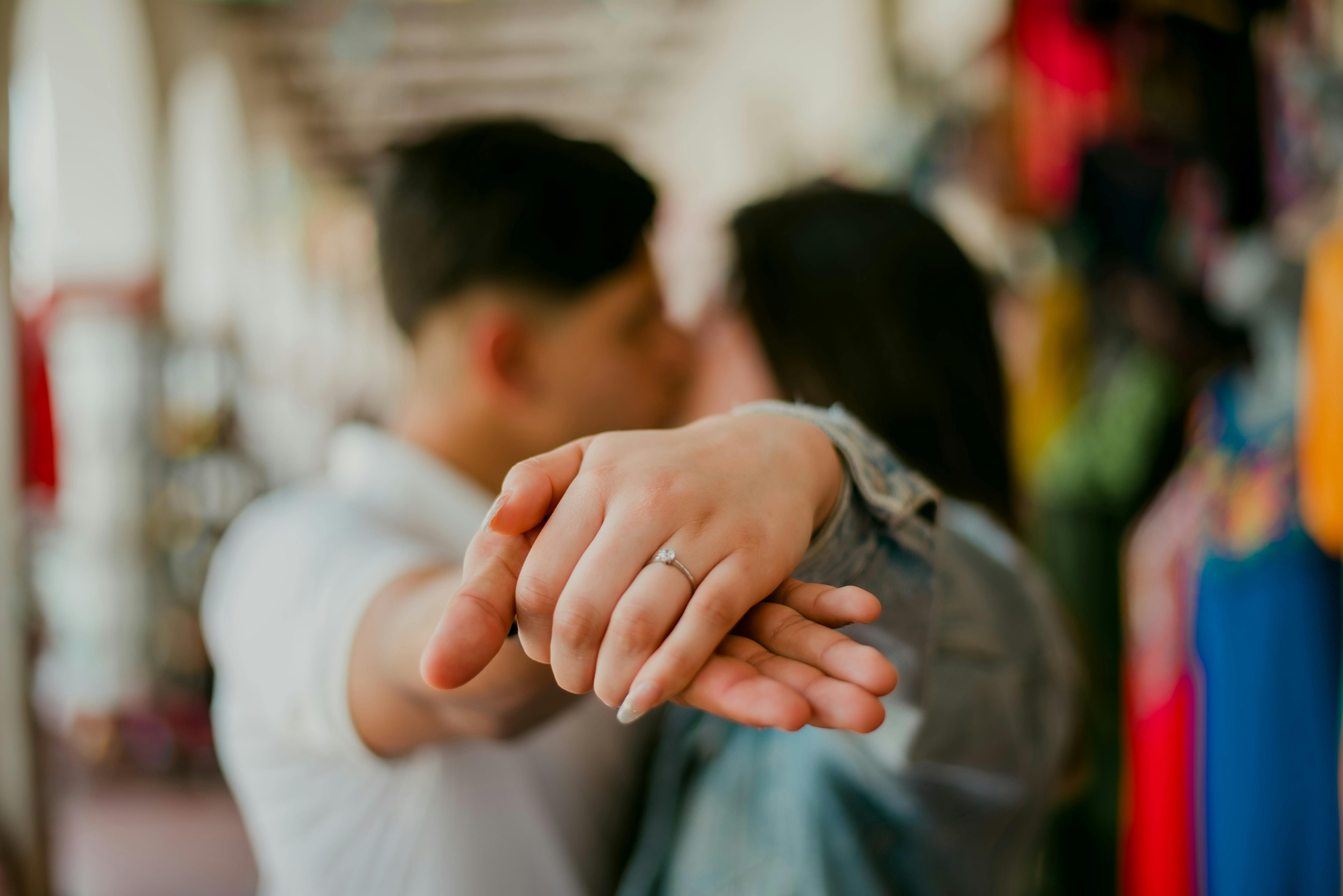 A couple shares a kiss as the focus is on their entwined hands showcasing an engagement ring.