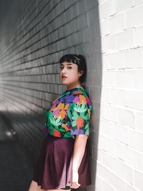A woman in a floral shirt and skirt leaning against a wall
