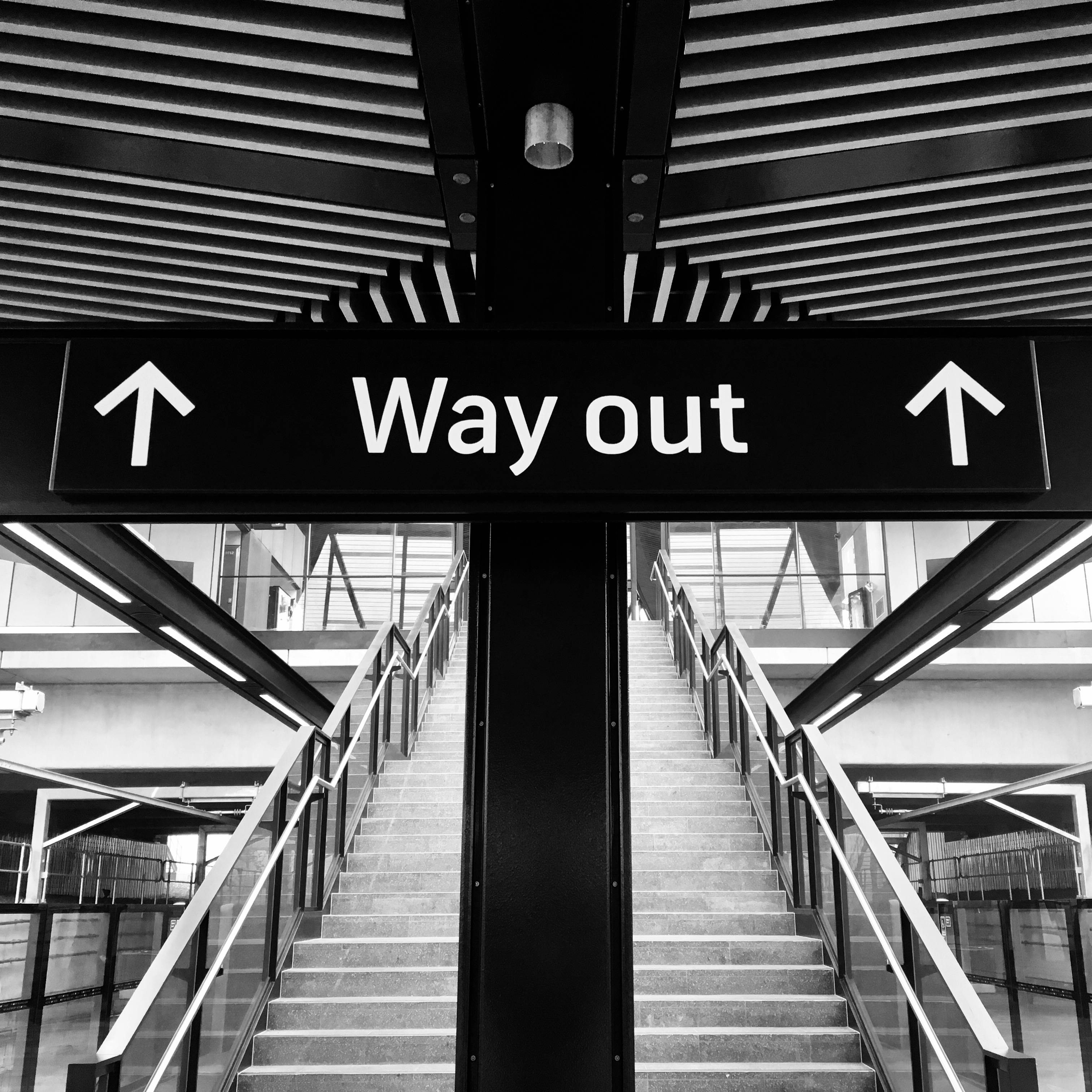 Monochrome image of a staircase with a 'Way Out' sign above, leading to an exit.