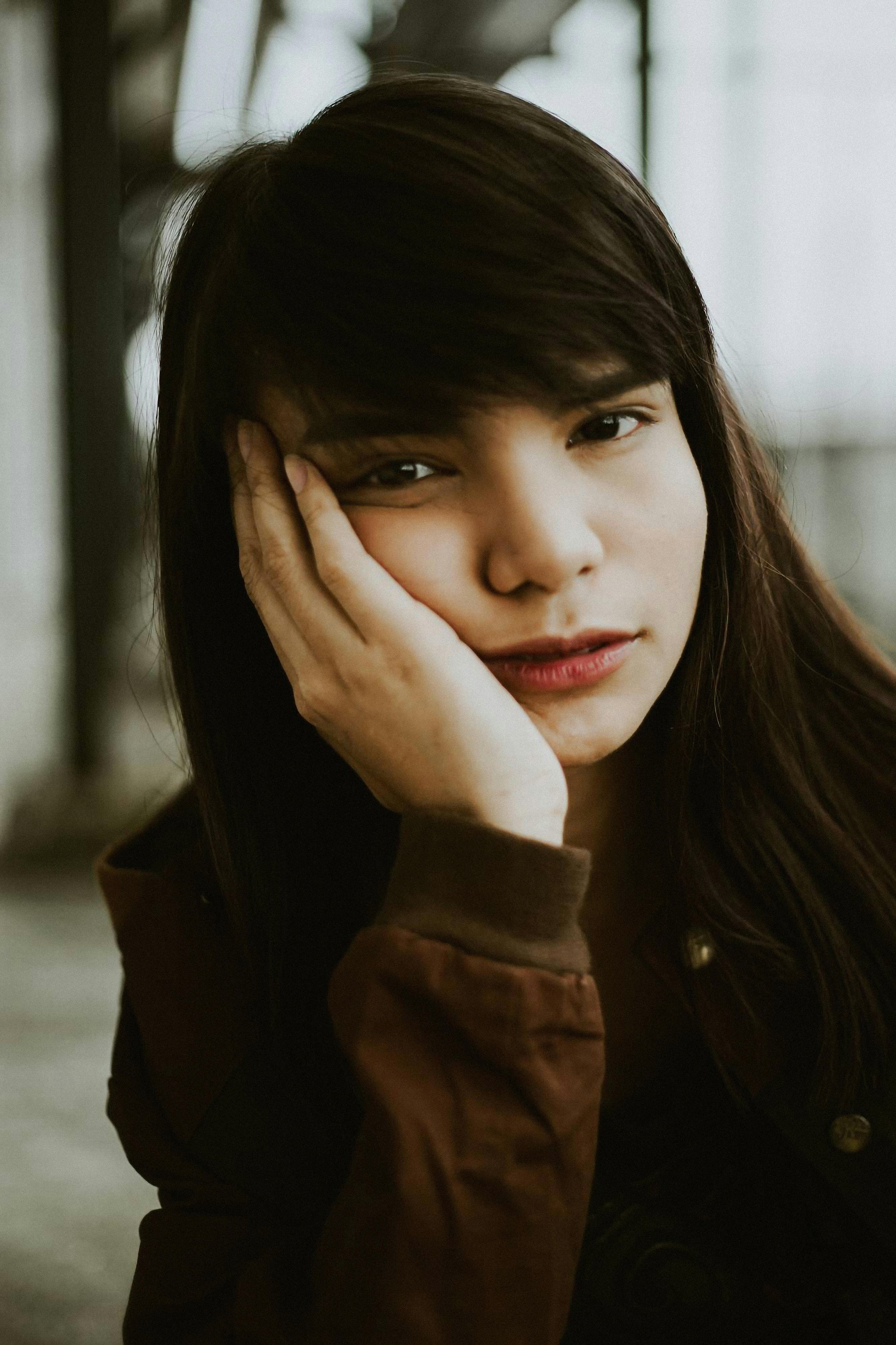 Portrait of a young brunette woman with bangs, resting her face on her hand, wearing a brown jacket.