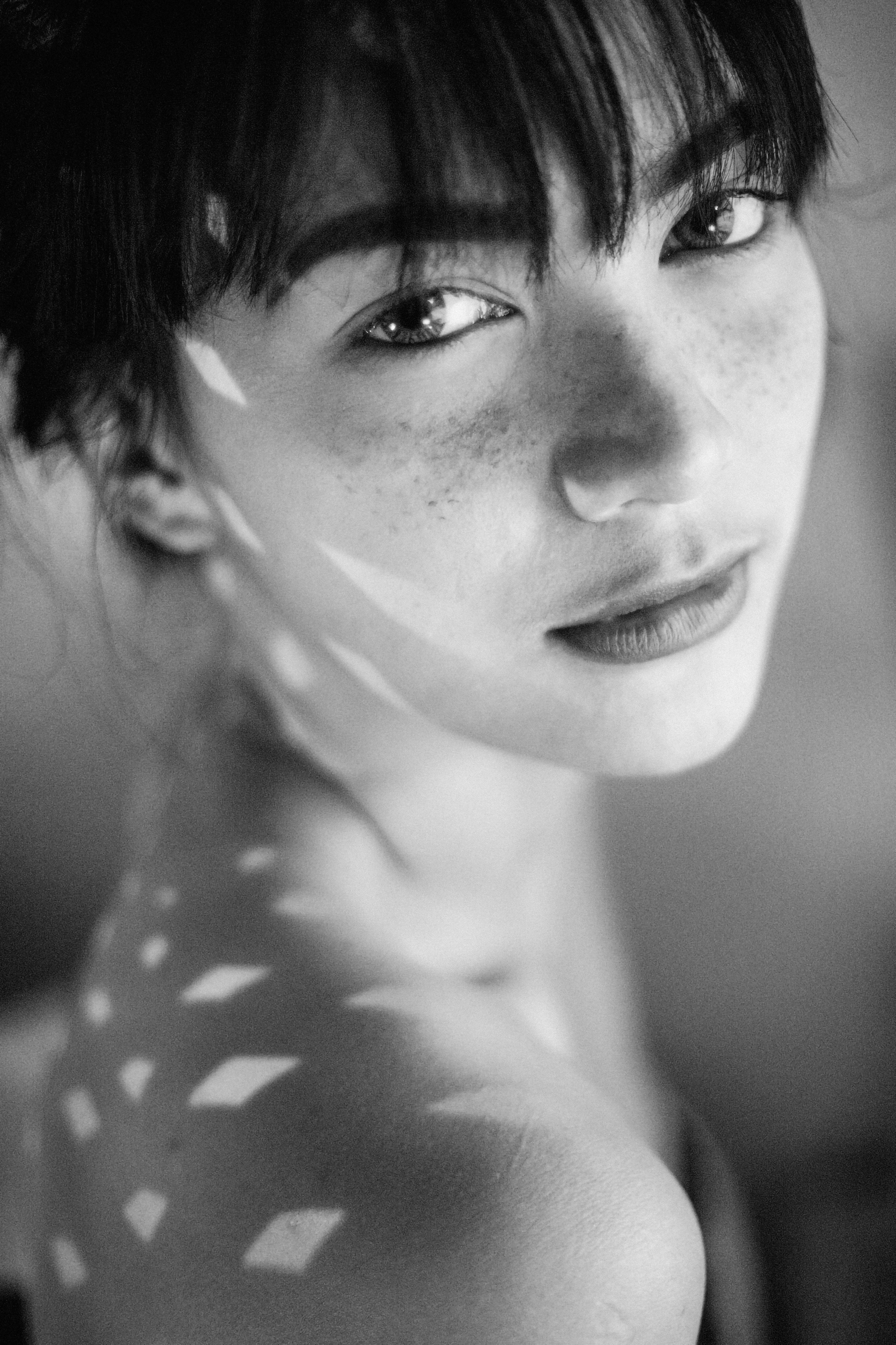 Striking black and white portrait of a young woman with freckles, illuminated by sunlight patterns.