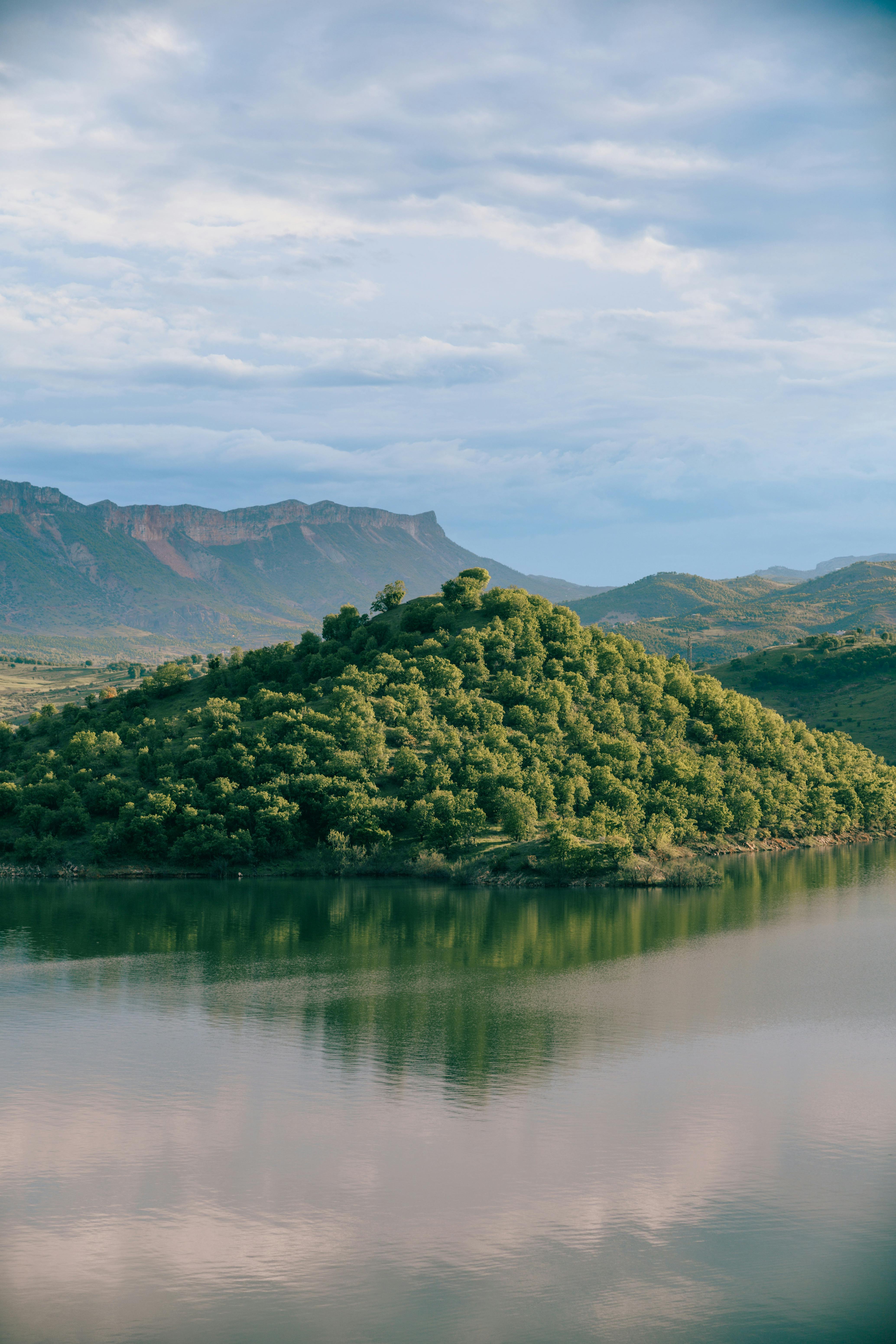 Green island reflected in a peaceful lake surrounded by mountains under a cloudy sky.
