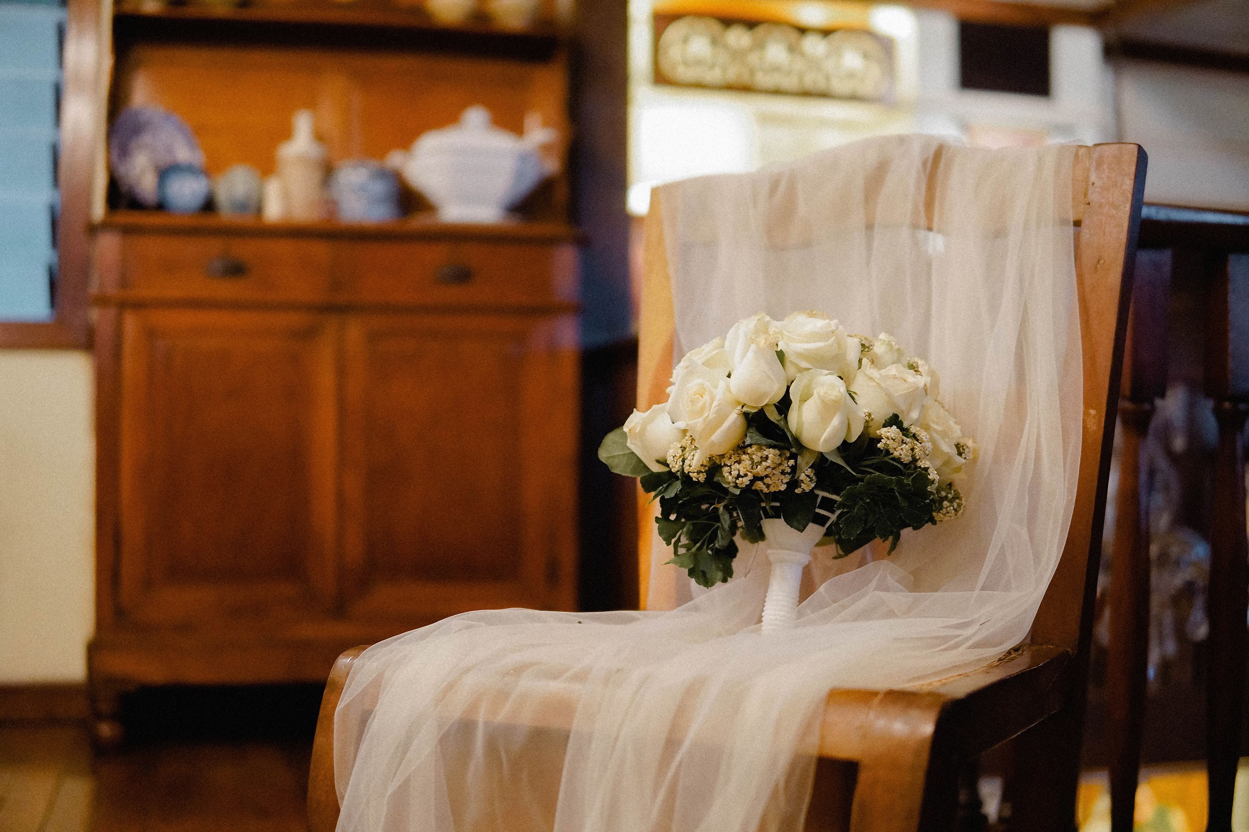 A wedding bouquet of white roses placed on a rustic wooden chair with a delicate veil, perfect for wedding themes.