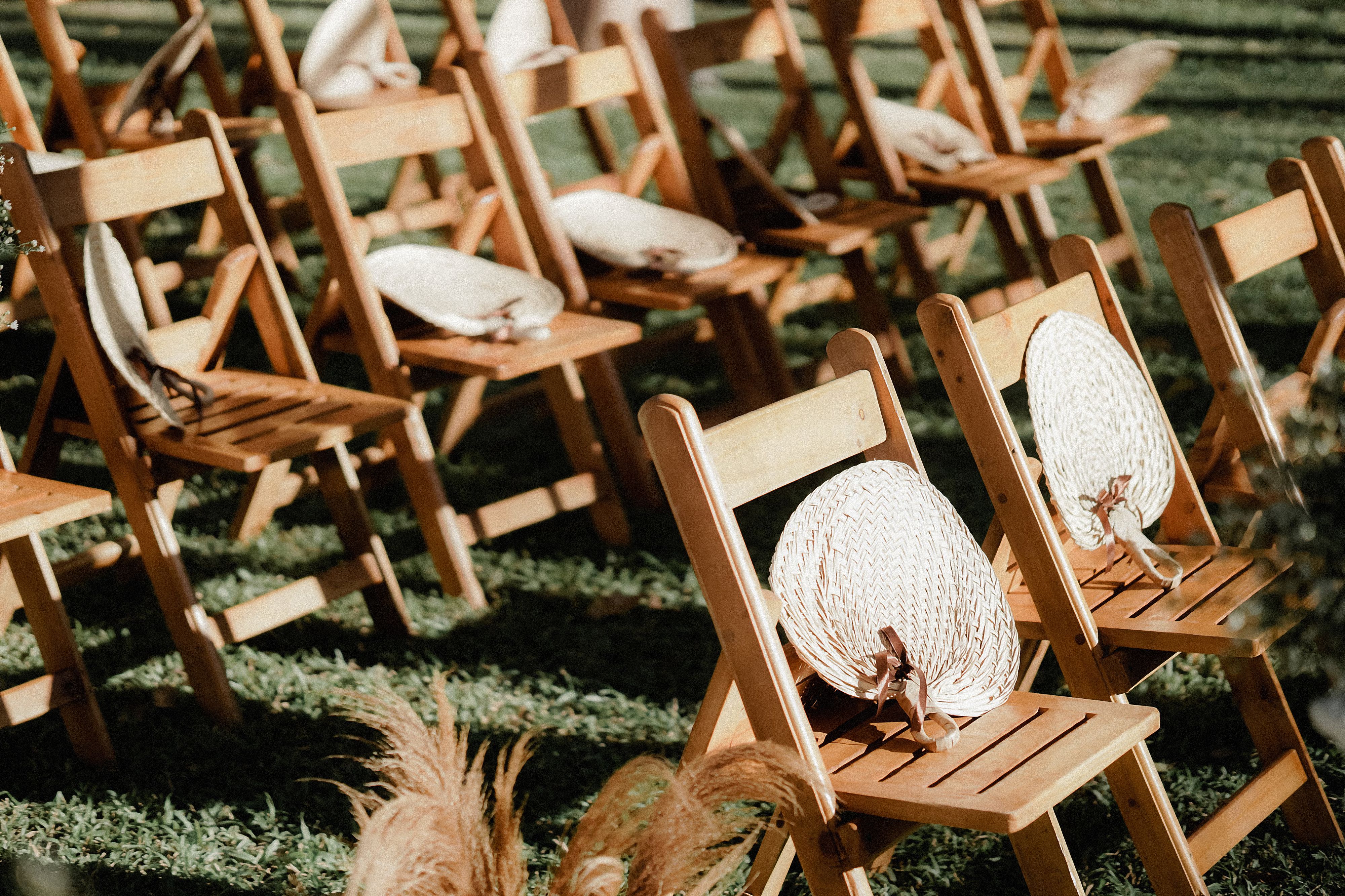 Outdoor wedding seating with wooden chairs and straw hand fans in a sunny garden setting.