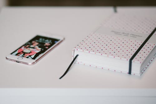 Free Rose Gold Iphone 6s Beside White and Black Polka Dots Book Both on Top of White Wooden Table Stock Photo