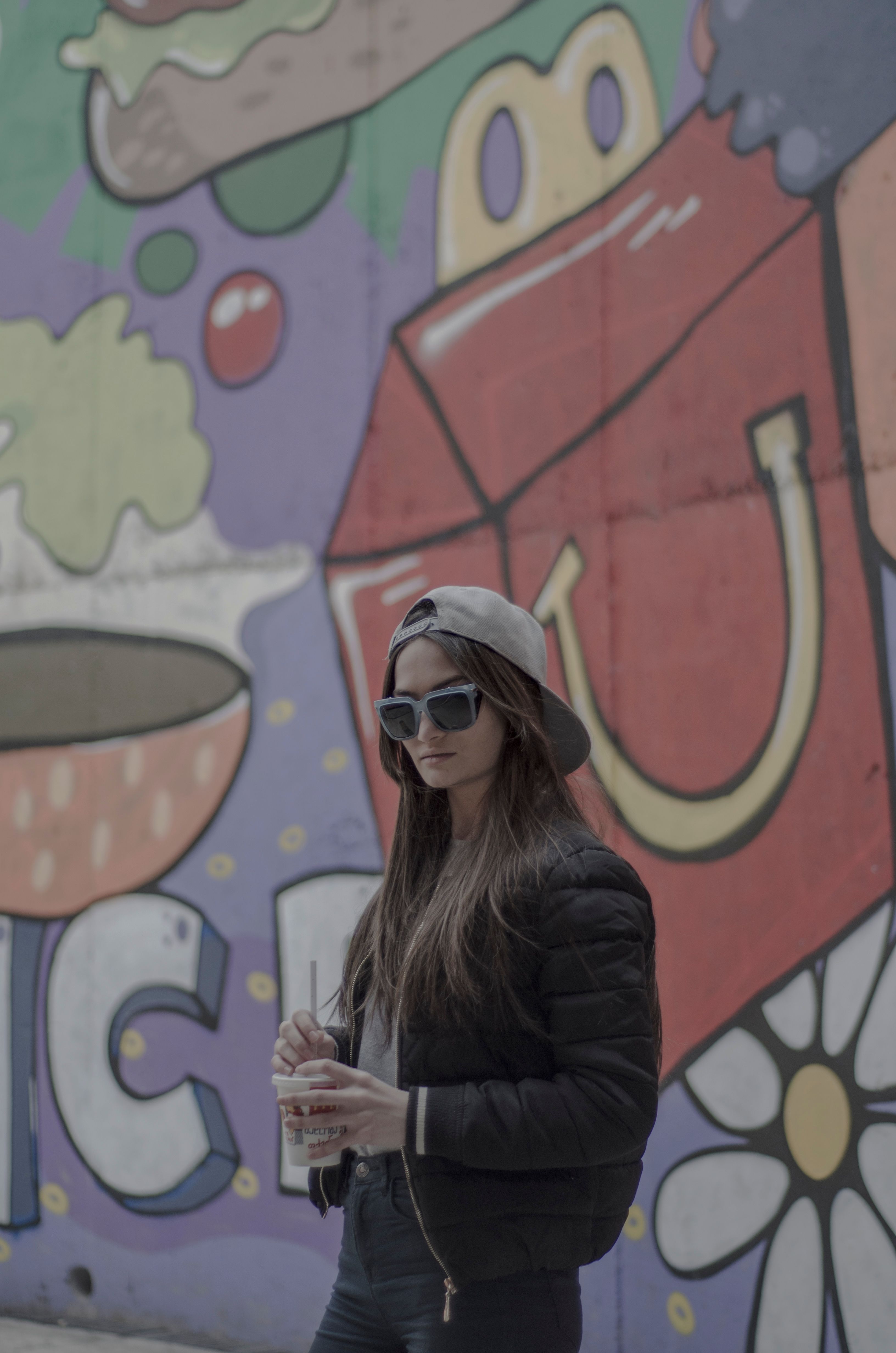 A stylish woman in sunglasses and cap poses in front of vibrant graffiti in Kutaisi, Georgia.