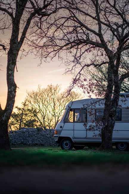 Self-contained motorhome parked on gravel beside a vineyard at sunset