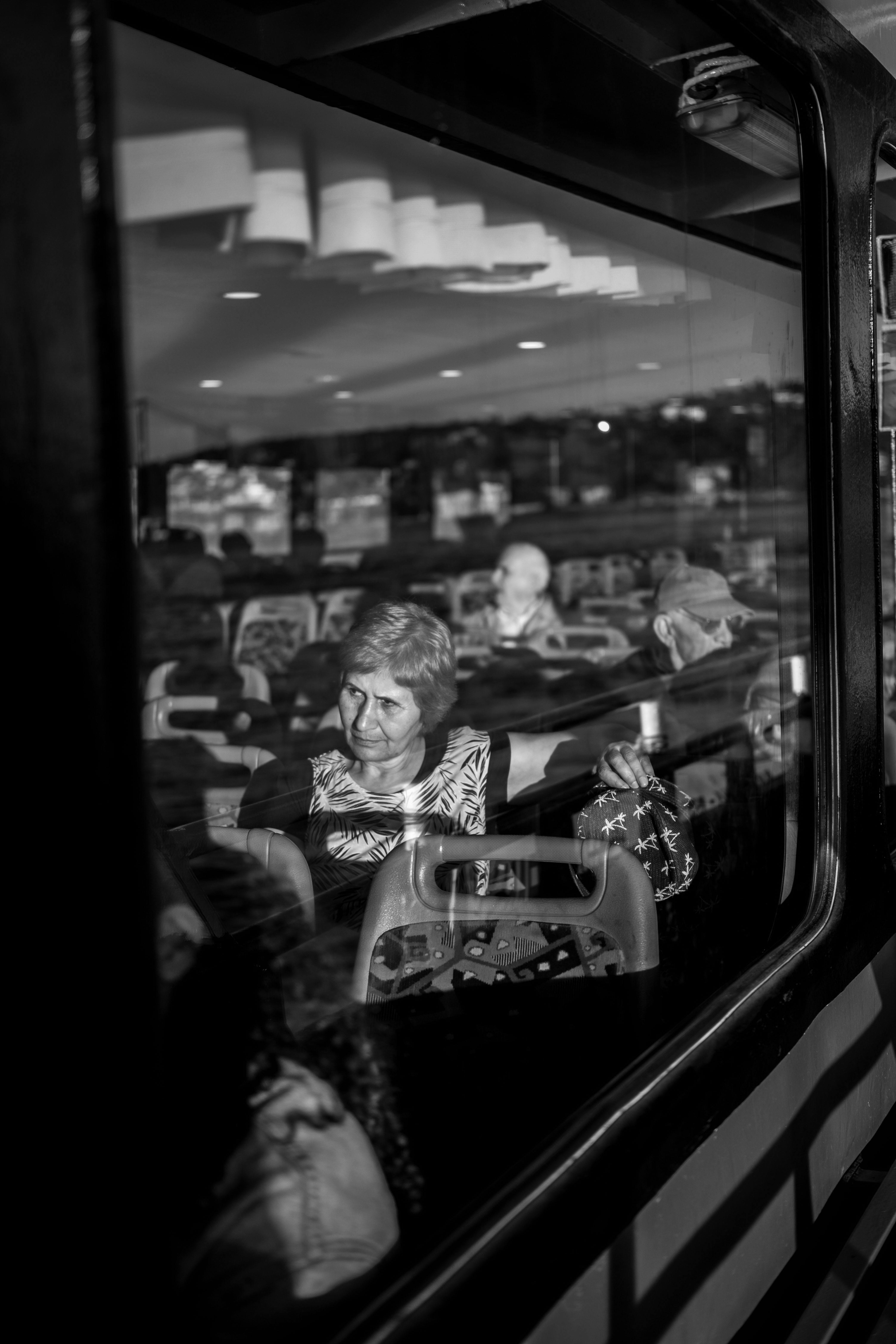 Black and white image capturing commuters on public transport, highlighting reflections and sunlight.