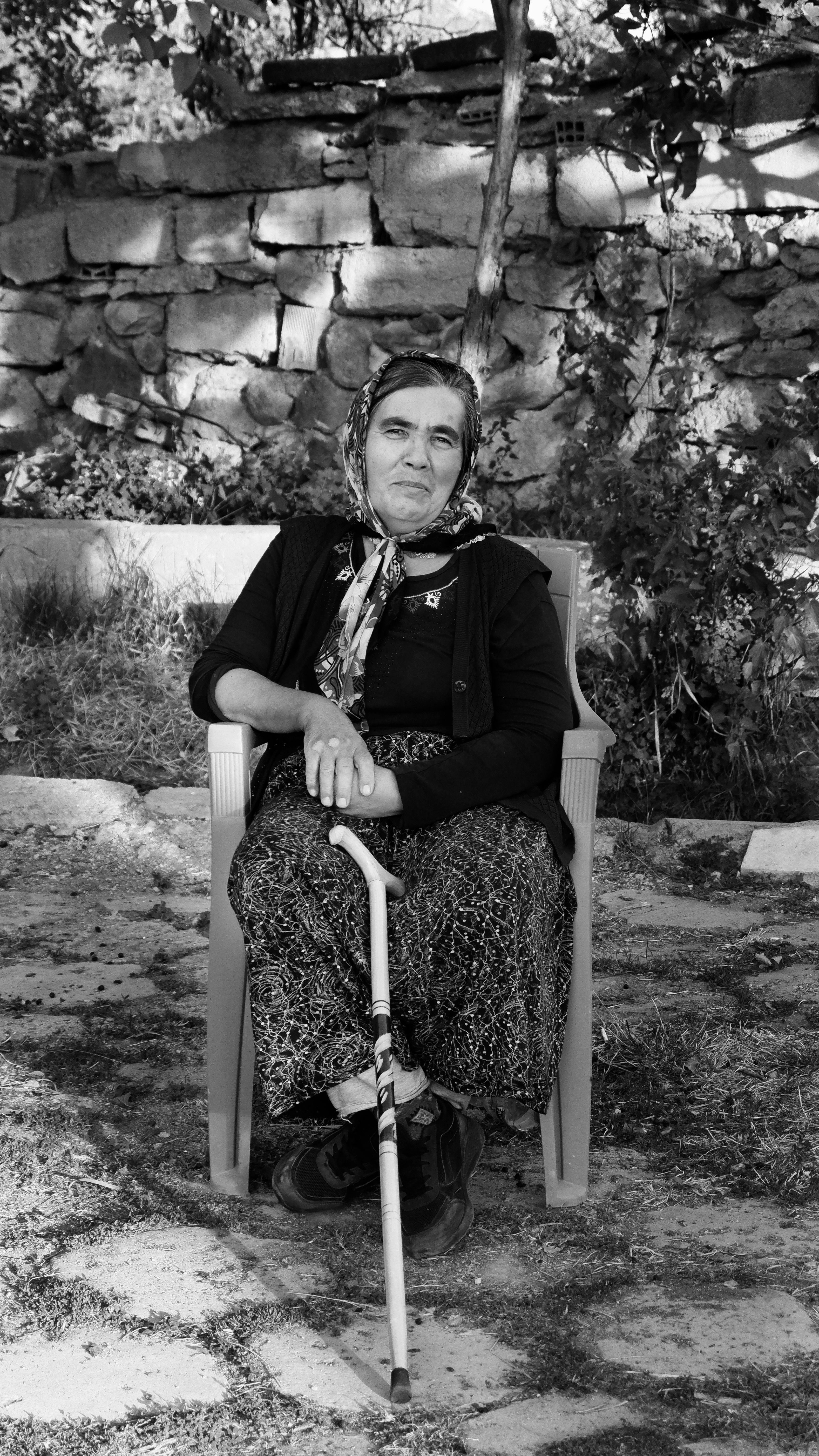 Elderly woman seated outdoors in a chair holding a walking stick, wearing a shawl.