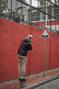 Man standing on bollard by a red wall and road sign, capturing urban exploration vibe.