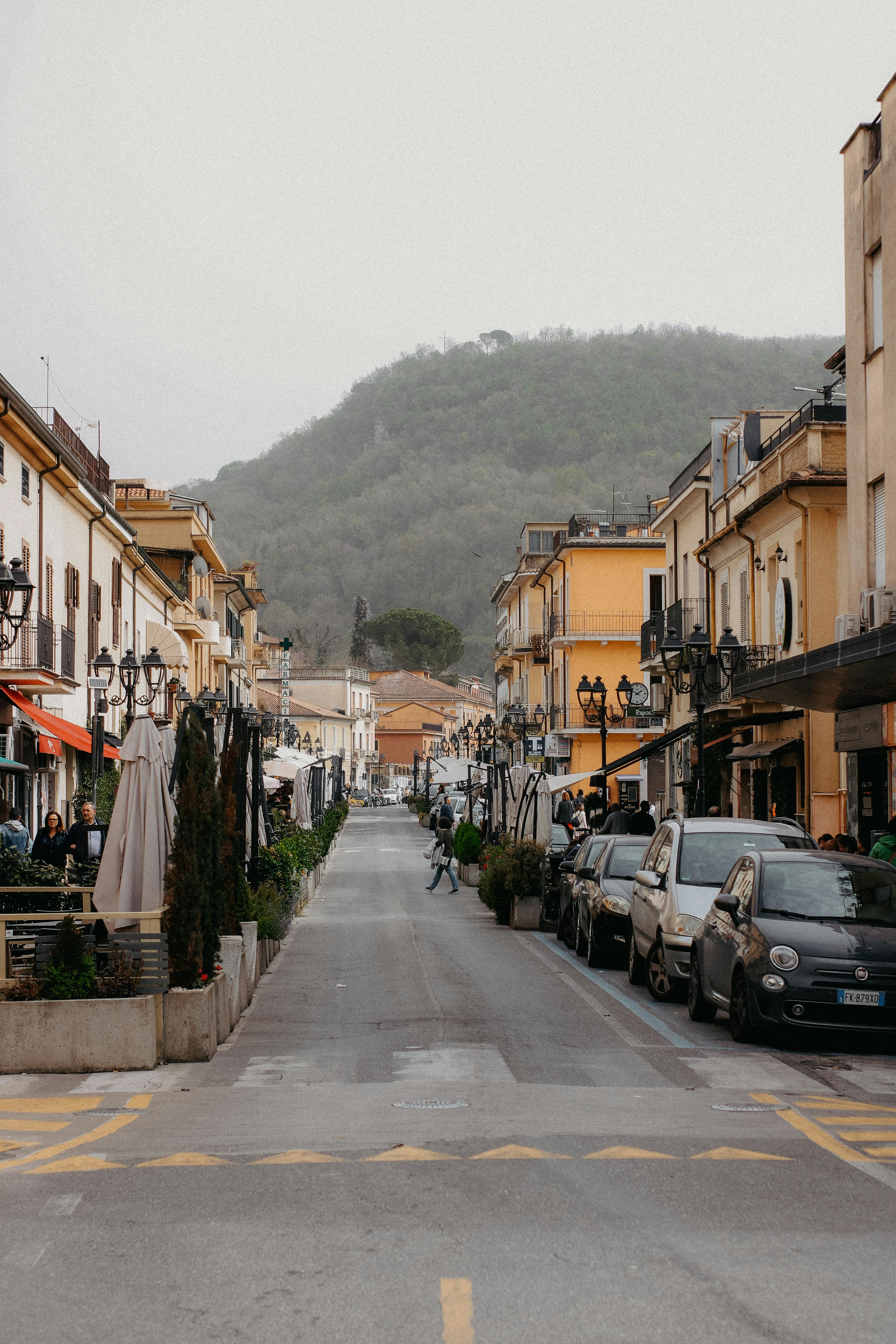 Capture of a picturesque urban street lined with cars and shops, framed by a lush hillside.