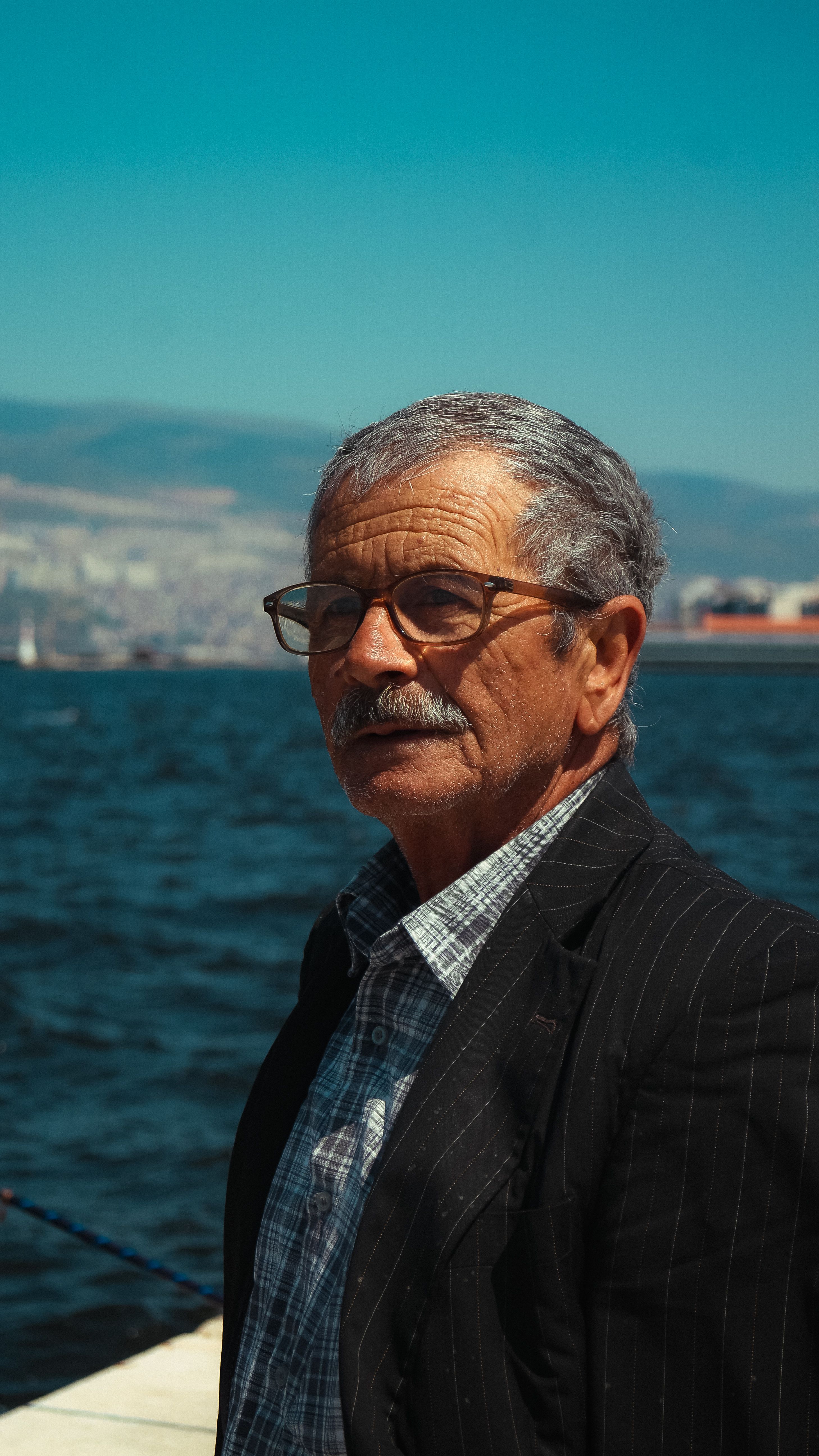 Senior man with glasses and a mustache standing by the sea under a clear blue sky.