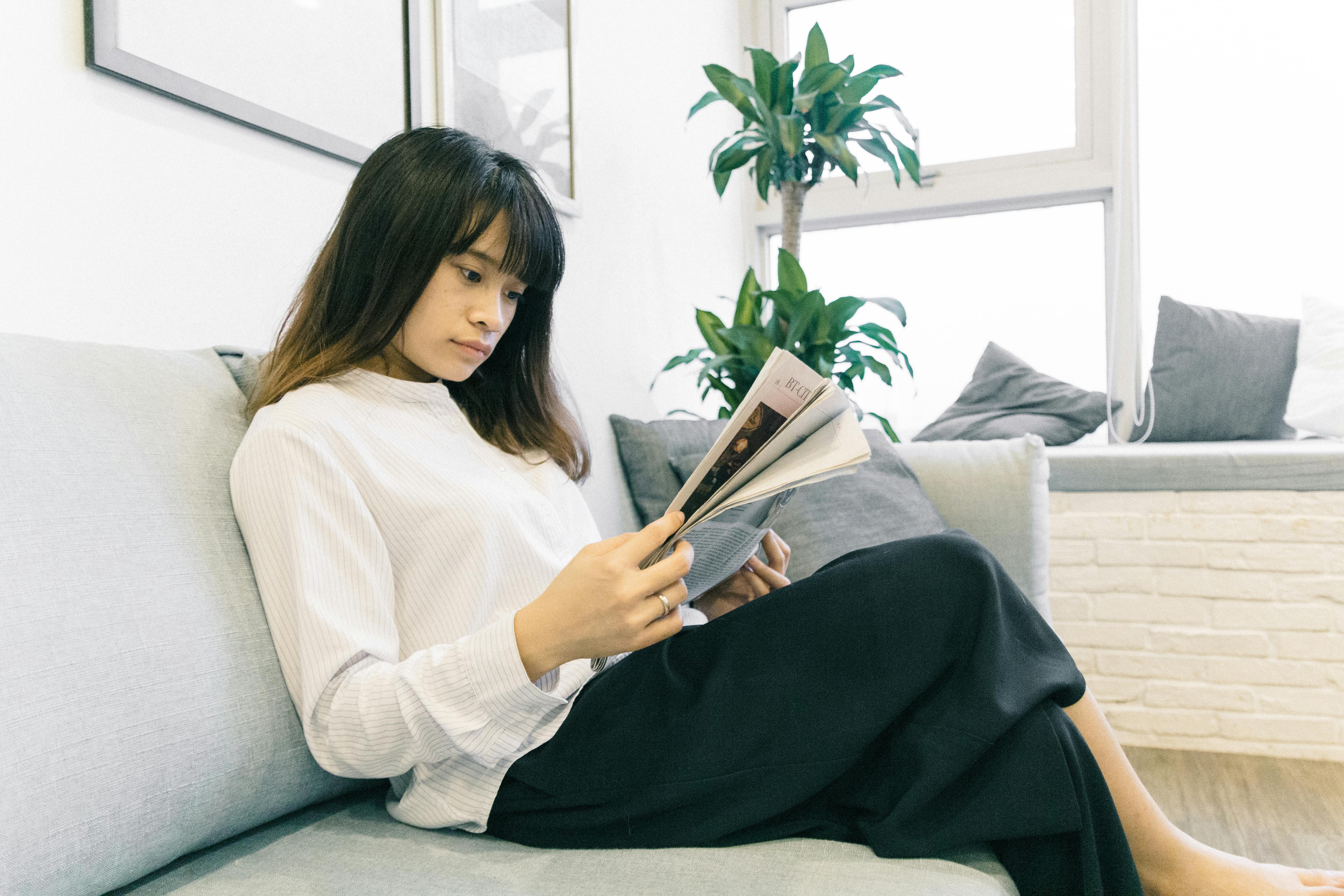 photo of woman reading magazine while sitting on gray sofa