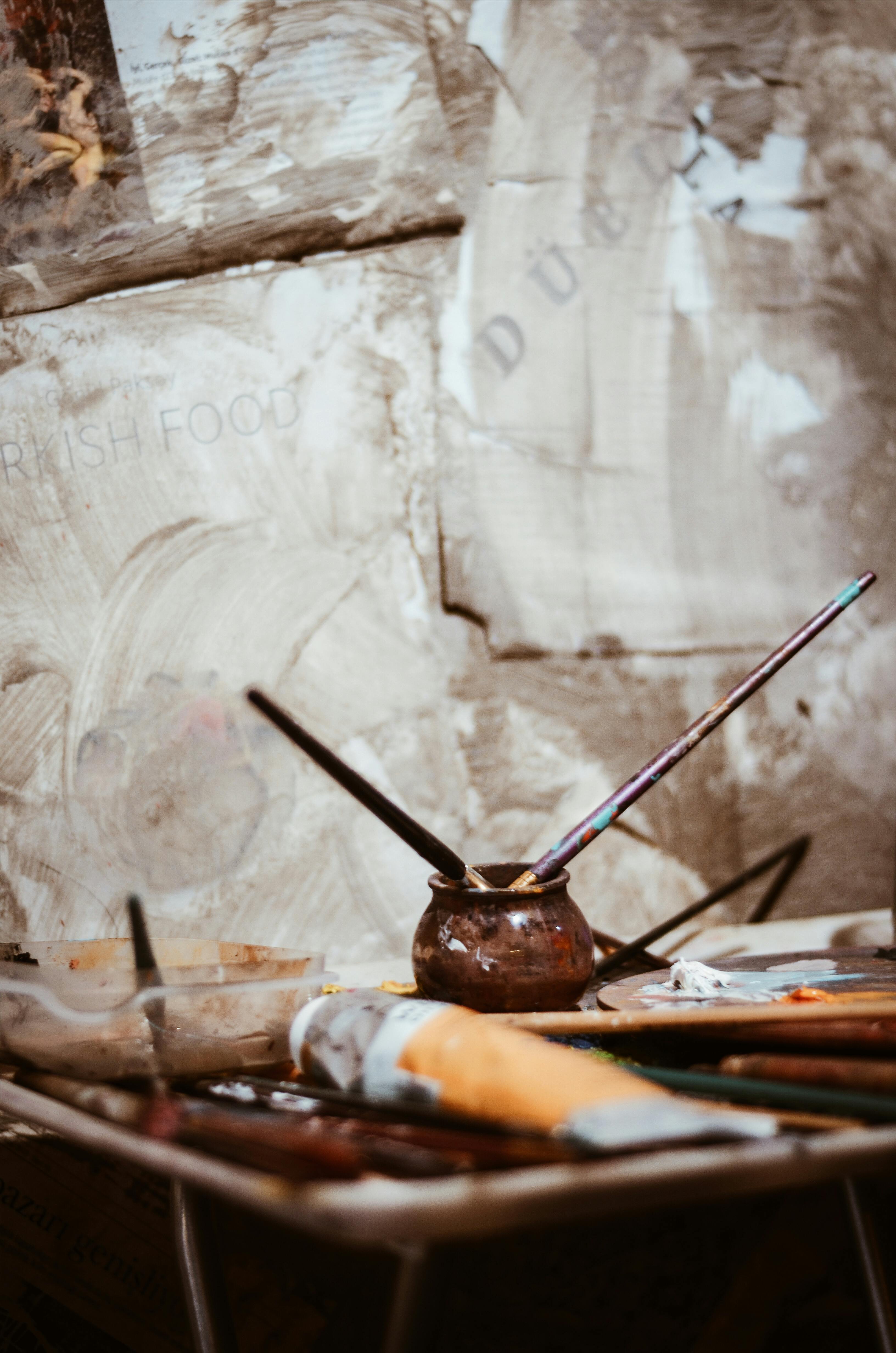 A pot of paint and brushes on a table in front of a wall.