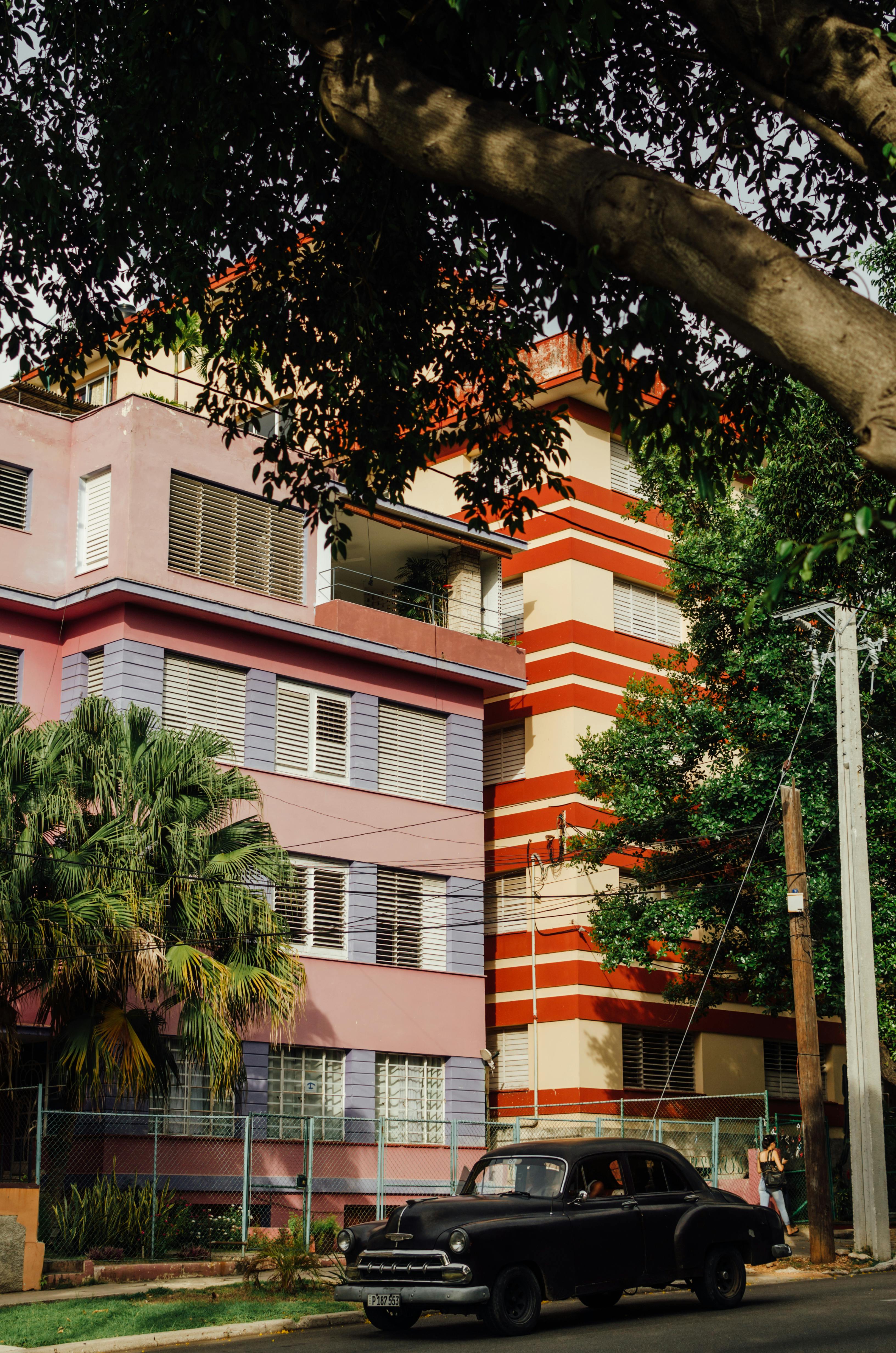 A vintage car parked on the street in front of colorful residential buildings in Havana, Cuba, with lush greenery.