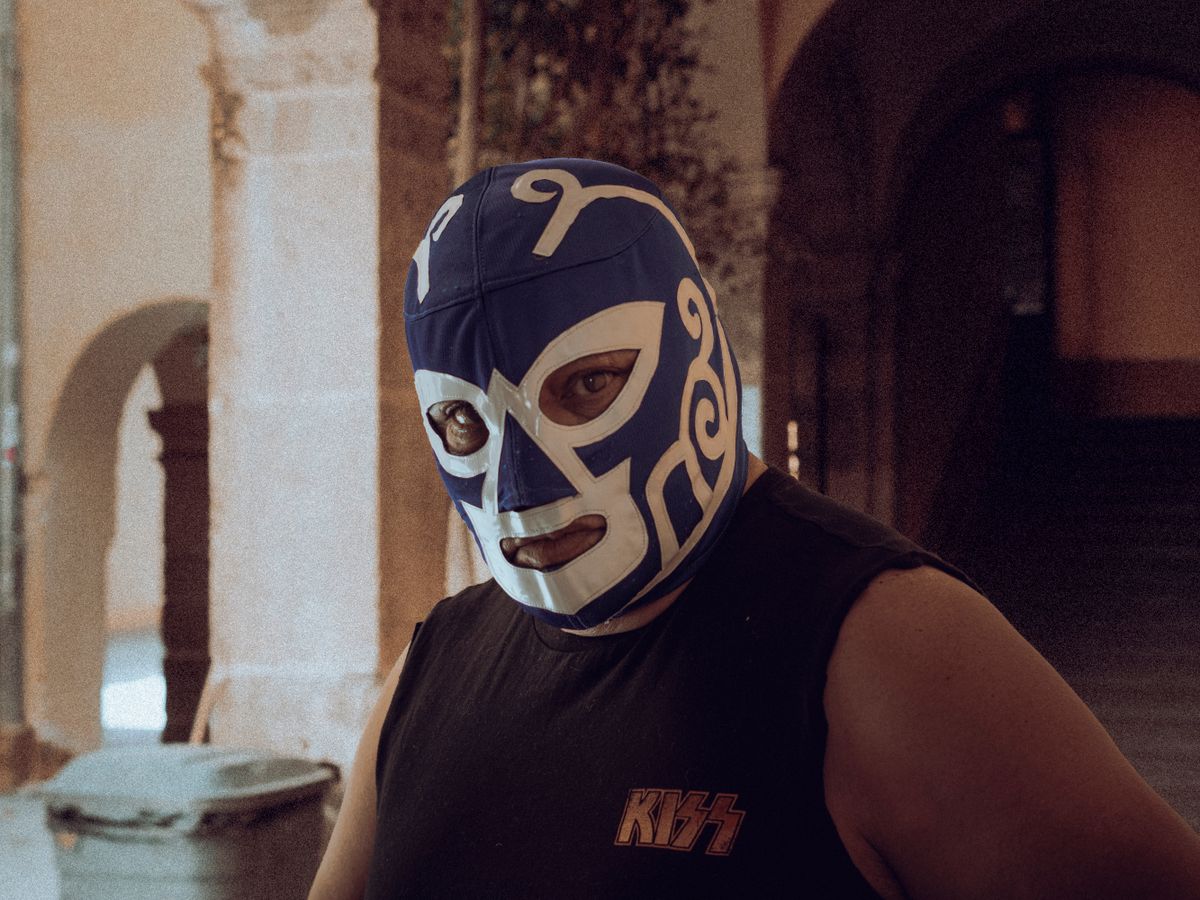 A luchador in a blue and white patterned mask standing in a colonnaded stone archway, a Kiss band shirt visible beneath his gear