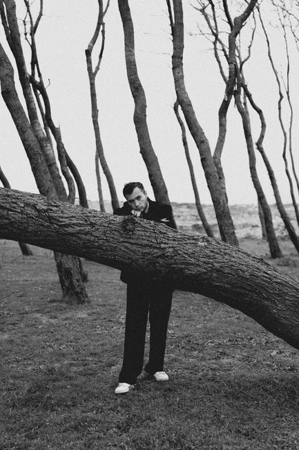 Black and white portrait of a man in a suit leaning on a bent tree in a forest.