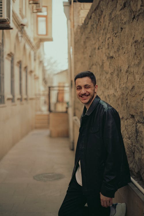 A man in a black jacket smiles in an alley