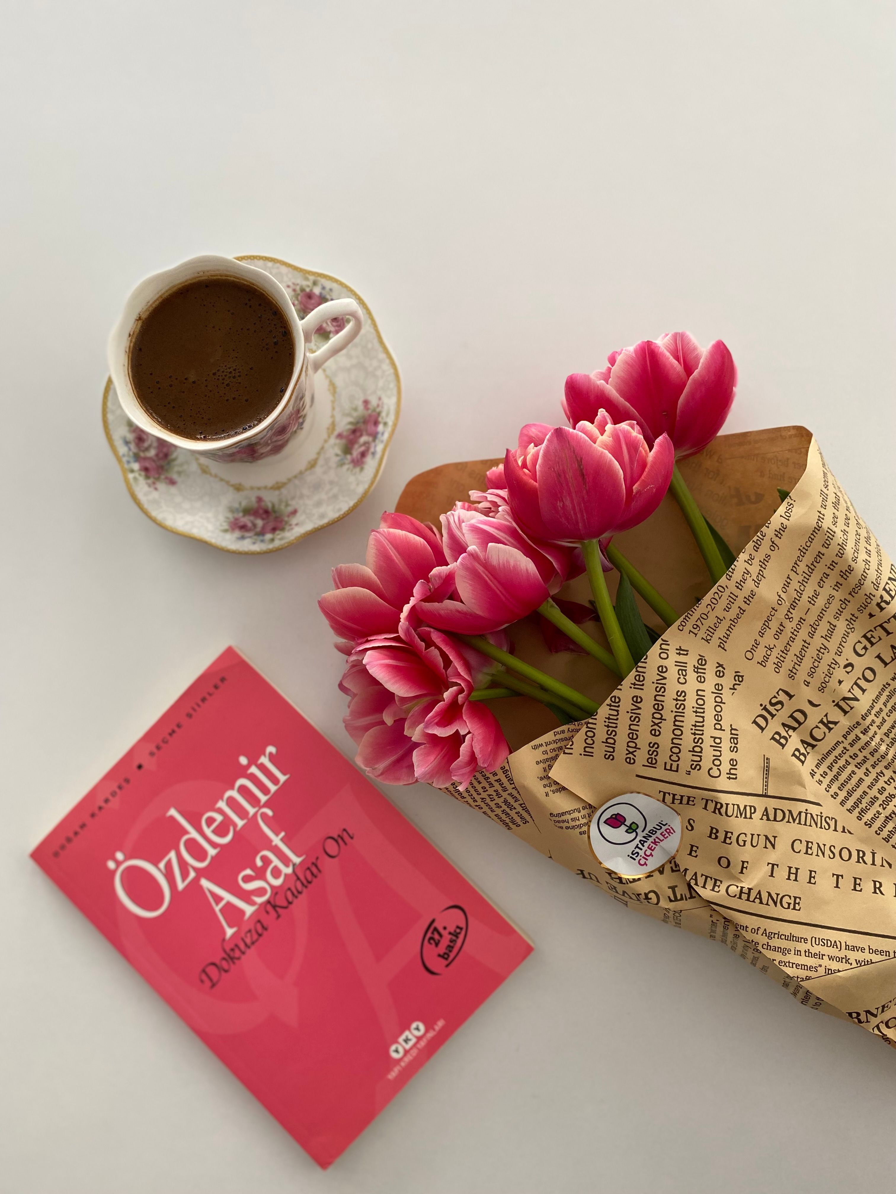 A still life composition featuring a cup of coffee, pink tulips, and a book on a white background.