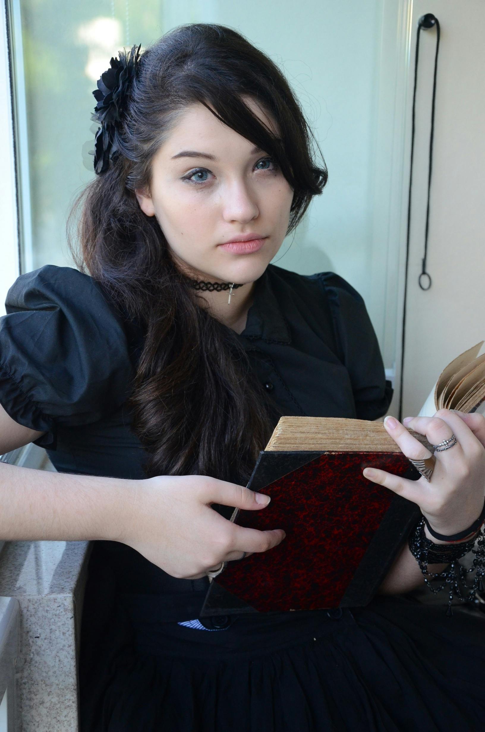 Free A young woman in Gothic attire reading a book by the window. Stock Photo