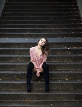 A stylish woman in a pink blouse and black boots sits on outdoor stairs, exuding confidence.