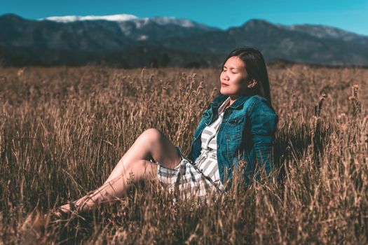 A peaceful Asian woman enjoying the sunshine in a wheat field with mountains in the background.