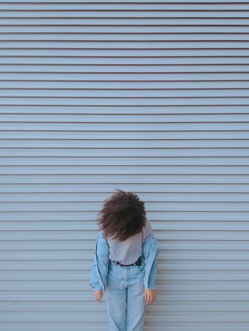 Young adult with curly hair in denim outfit against pastel wall in urban setting.