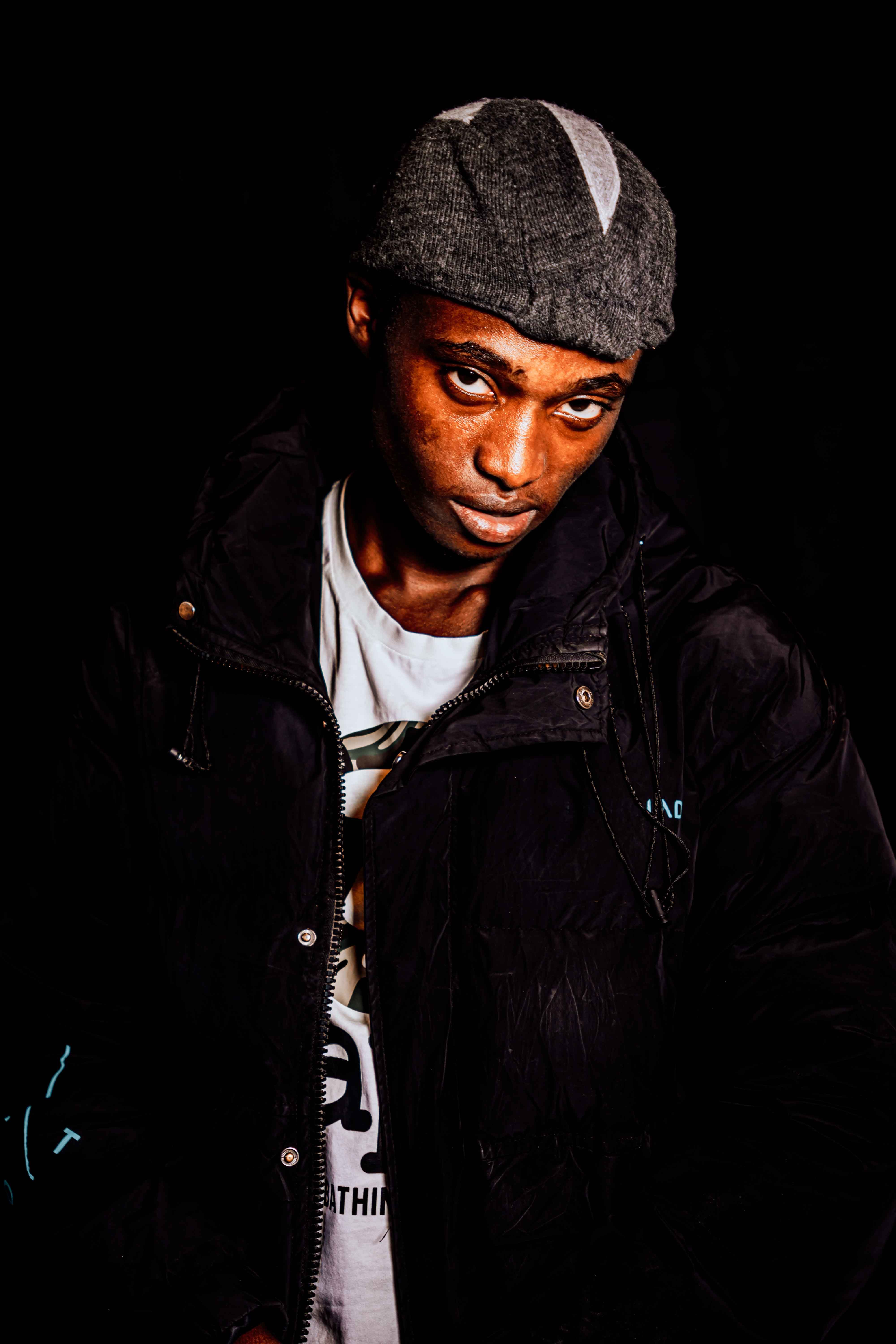 Portrait of a man wearing a black jacket and cap against a dark background, capturing urban fashion vibes.