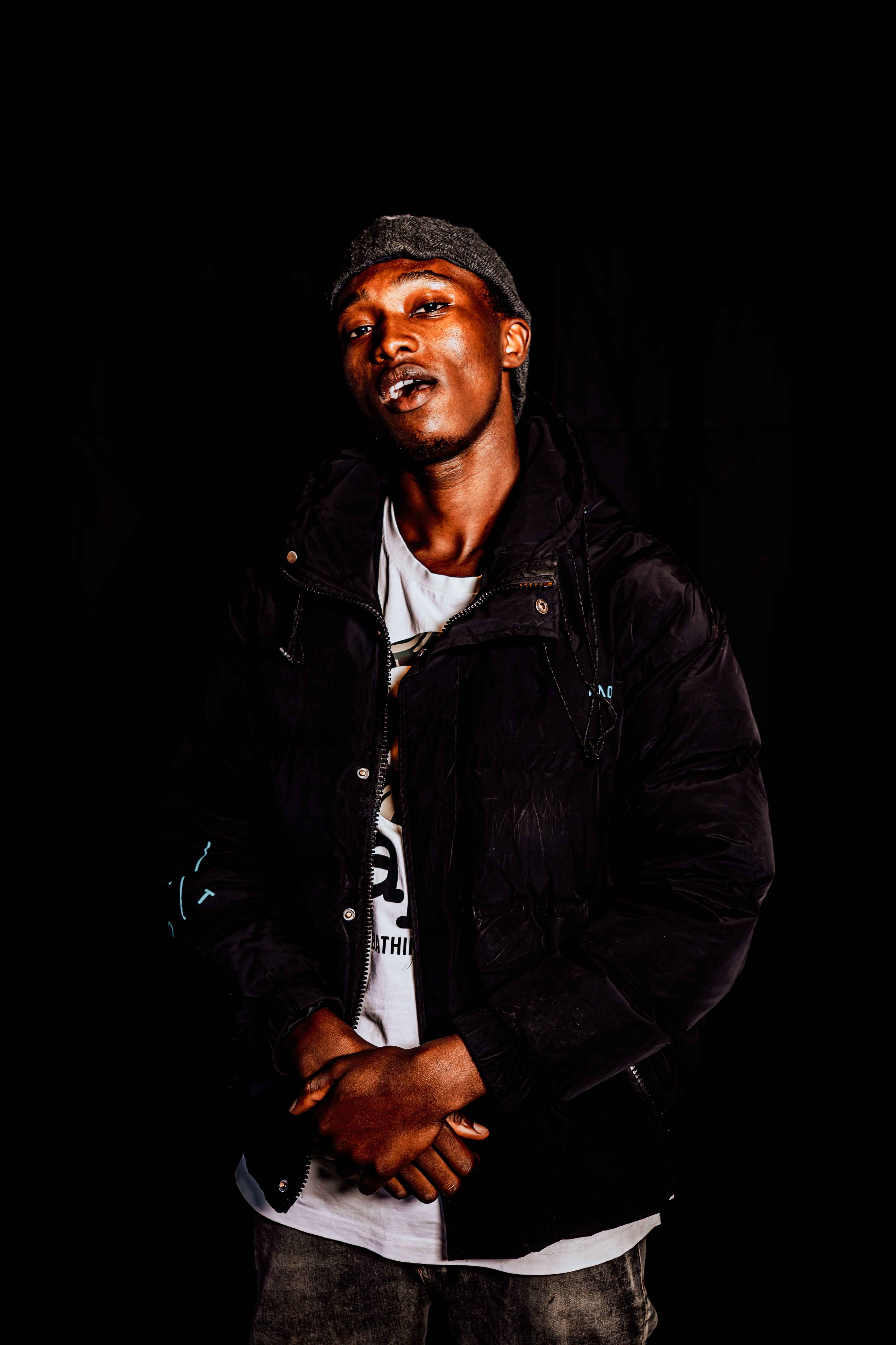 A fashionable young man in a black jacket and cap posing against a dark background.