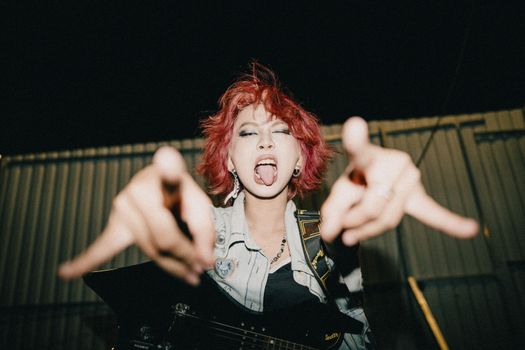 Dynamic photo of a female rocker with red hair performing and posing with an electric guitar outdoors at night.