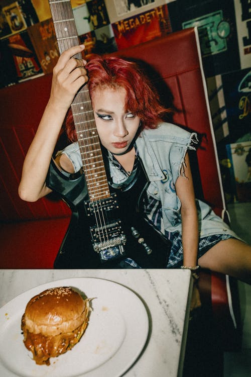 A woman with red hair holding a guitar and a burger