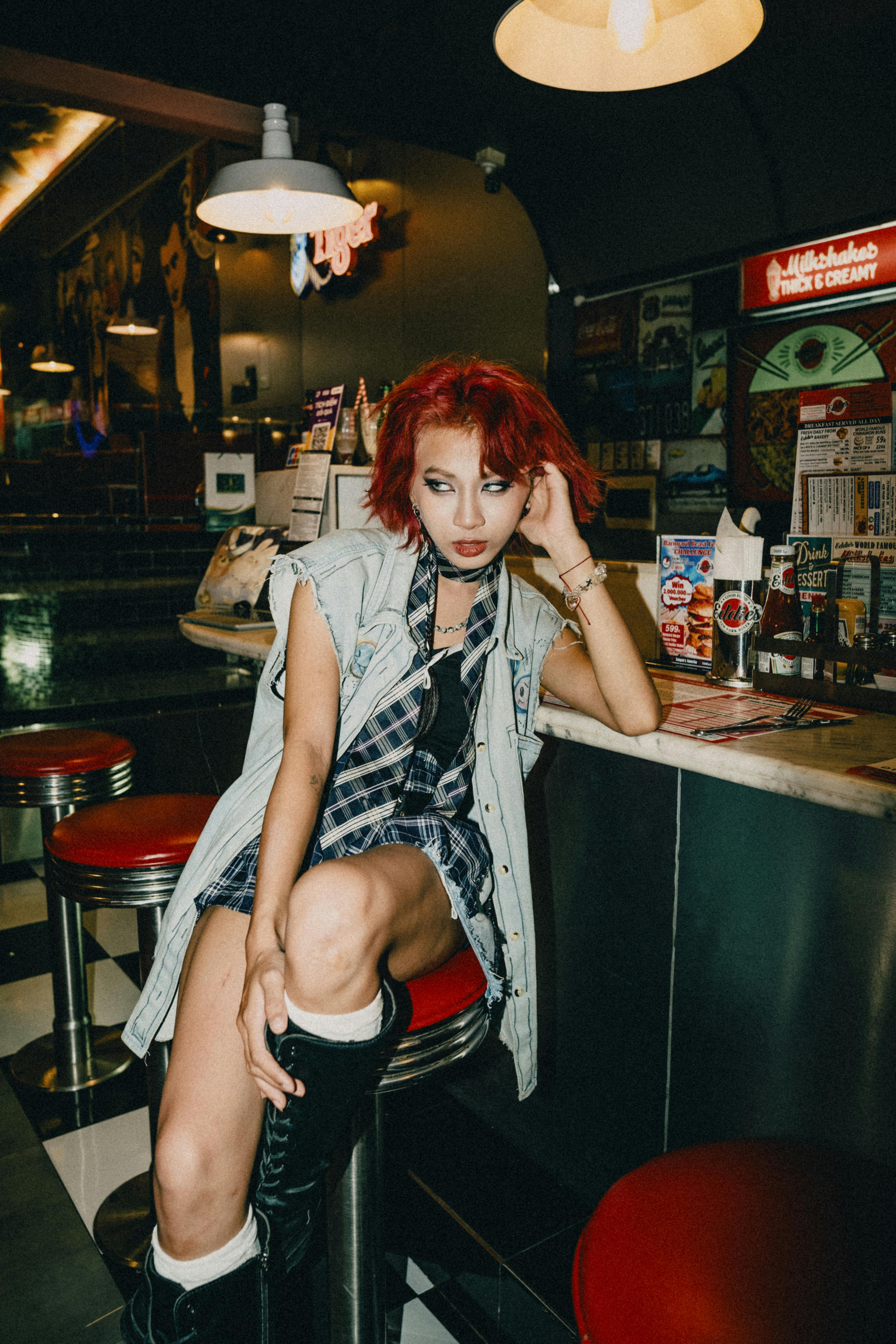 Stylish woman with dyed red hair sitting at a retro diner bar counter, showcasing edgy fashion.