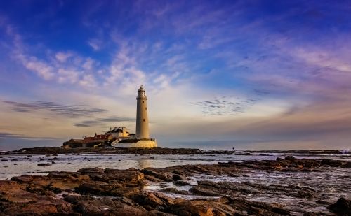 Free Lighthouse overlooking the rocky seashore at dusk, under a vibrant sky. Stock Photo